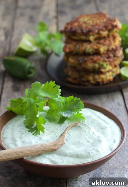 A close-up of zesty Cilantro-Lime Sour Cream in a white bowl with a wooden spoon, with blurred zucchini fritters in the background.