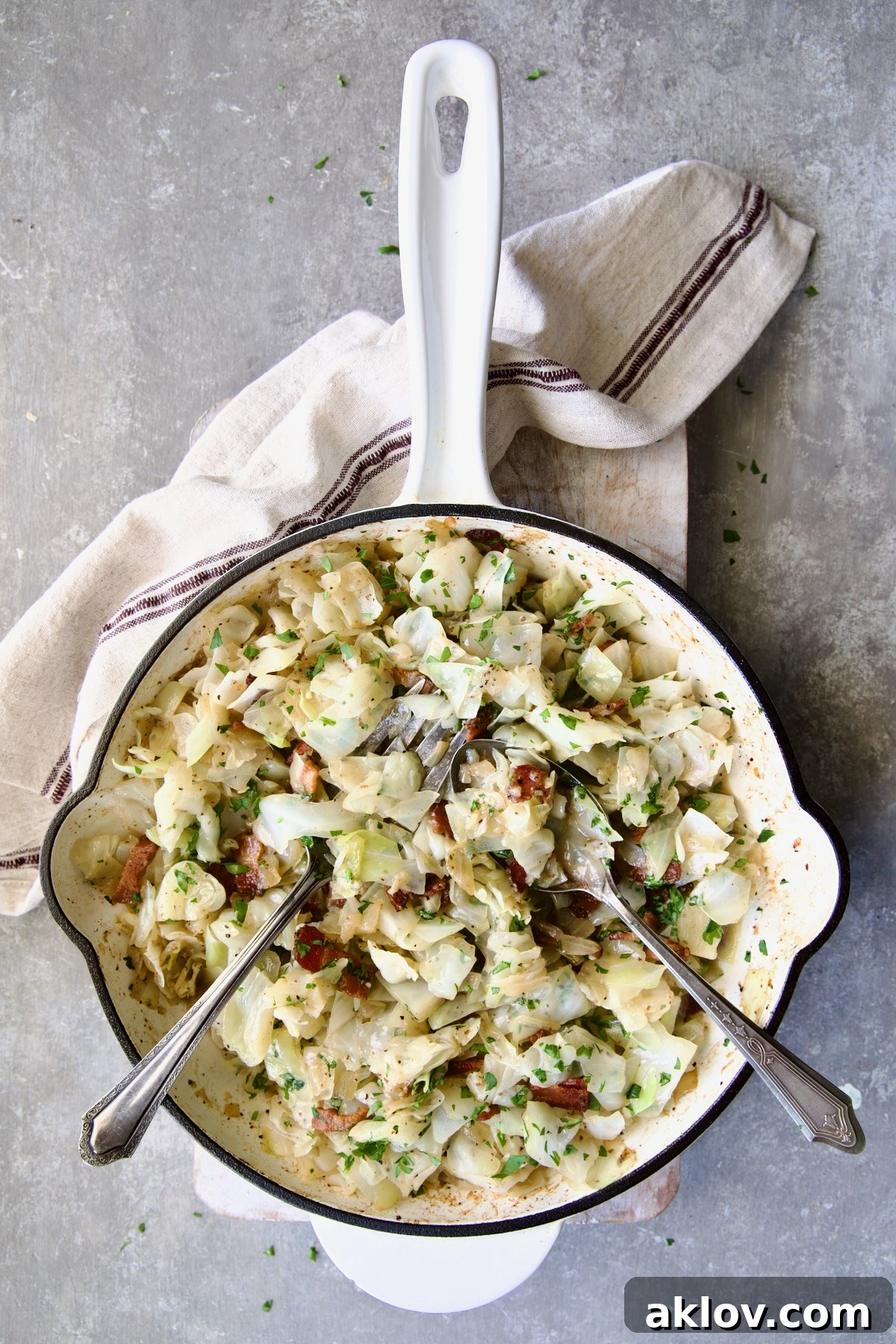 Fried Cabbage with Bacon in white enamel cast iron skillet with serving utensils on white cutting board with red striped napkin.