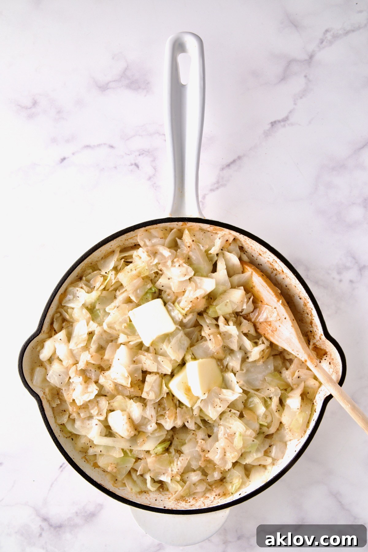 Butter being stirred into cabbage in white enamel cast iron skillet with wooden spoon.