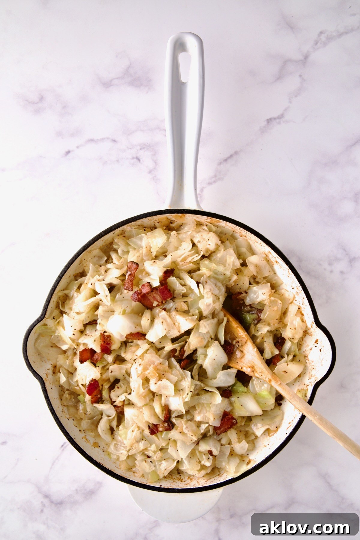 Bacon being stirred back into cabbage in white enamel cast iron skillet.