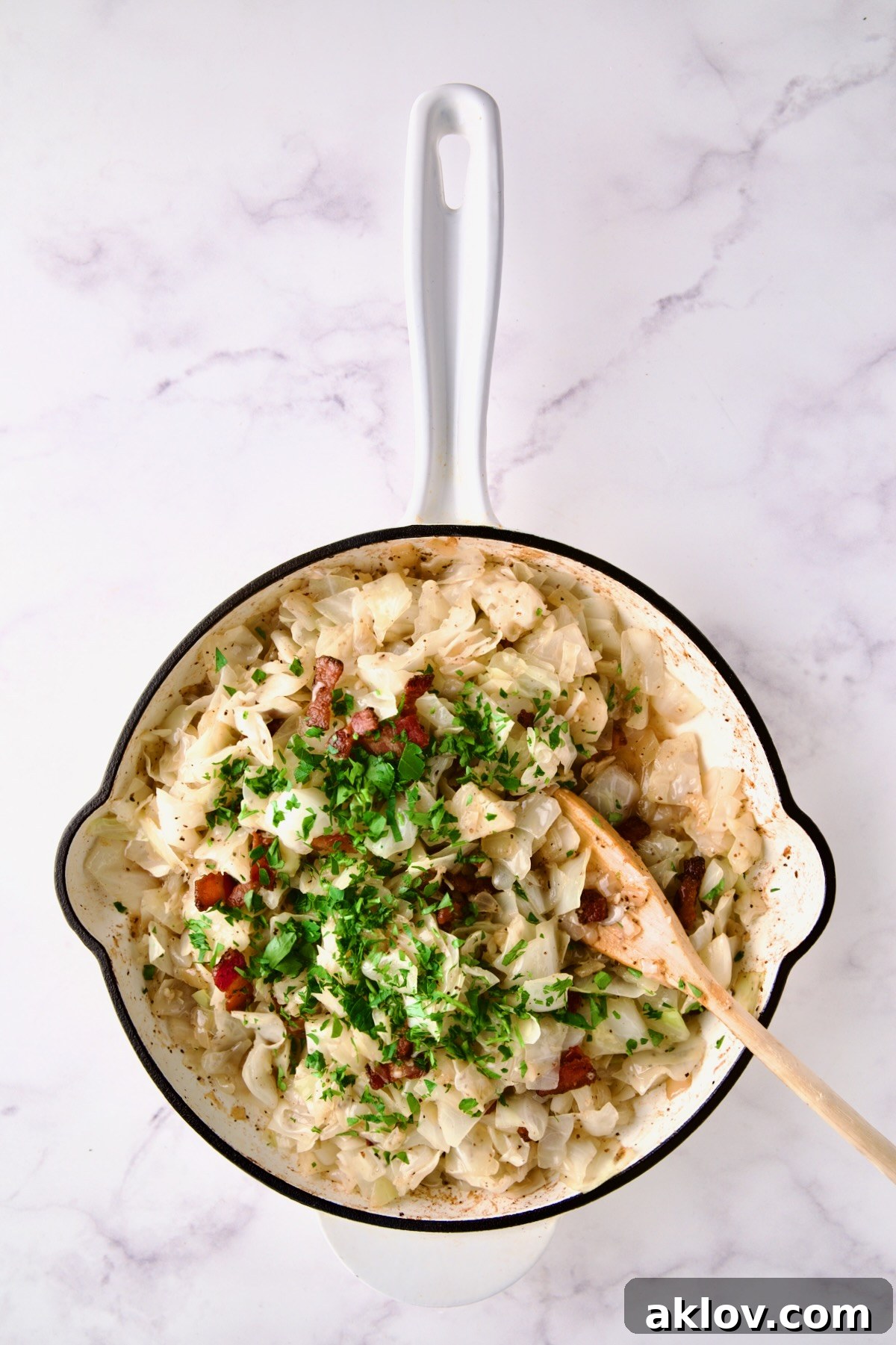 Cabbage in white enamel cast iron skillet with bacon and parsley added being stirred with wooden spoon.