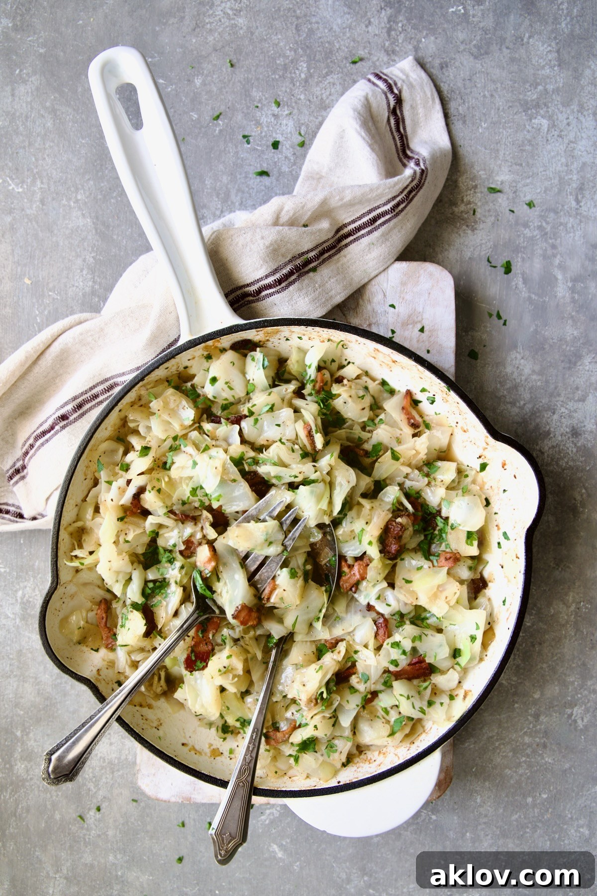 Fried Cabbage with Bacon in white enamel cast iron skillet with serving utensils and red striped napkin.