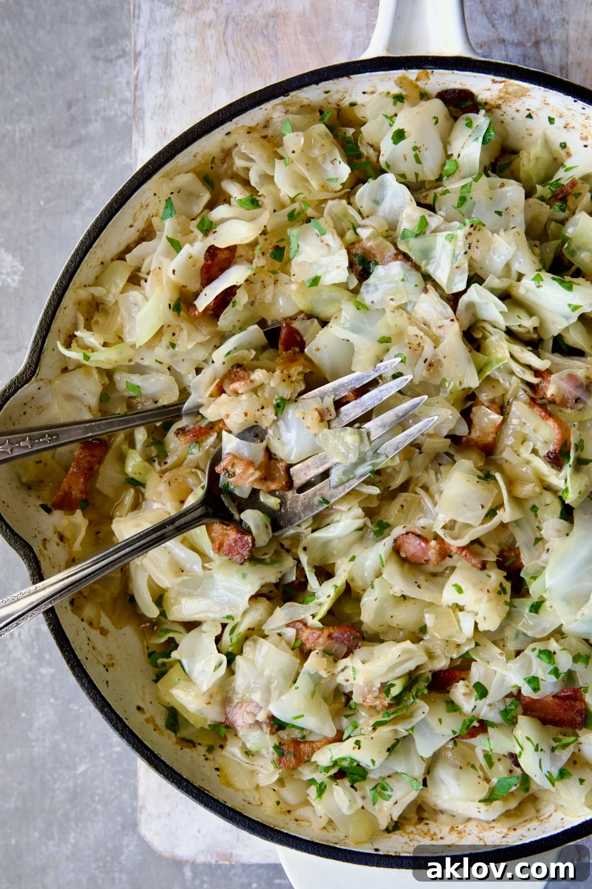 Two serving utensils serving up Fried Cabbage with Bacon from white enamel cast iron skillet.