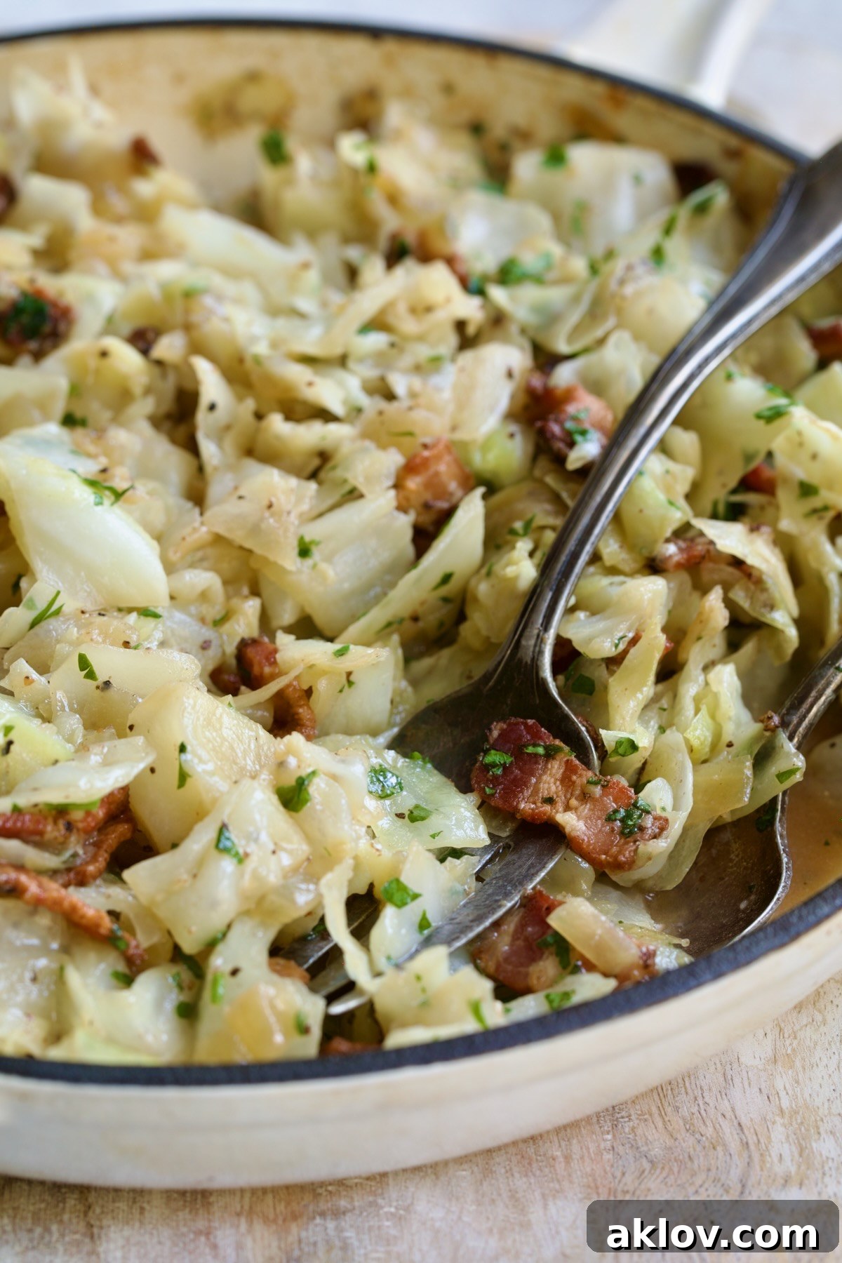 Finished Fried Cabbage with Bacon in white enamel cast iron skillet with serving utensils.
