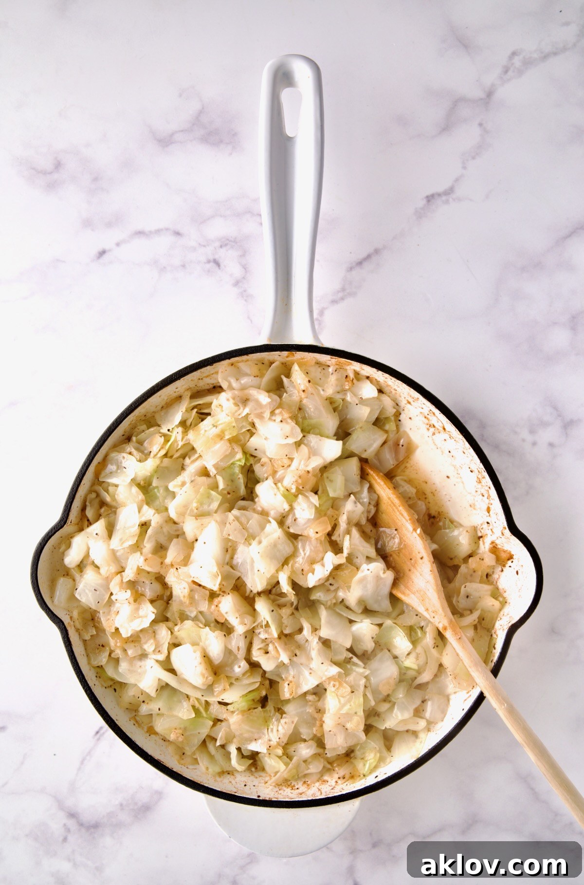 Wooden spoon stirring cabbage after chicken broth is added in white enamel cast iron skillet.