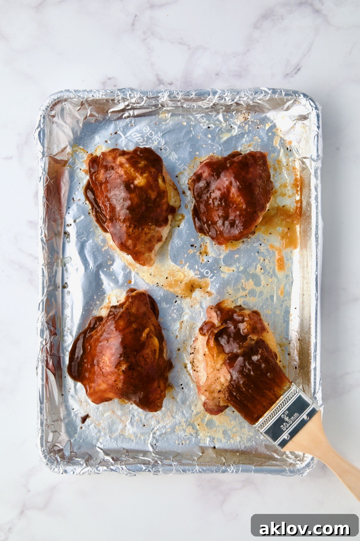 Four chicken thighs being brushed with barbecue sauce on aluminum foil-lined baking sheet.