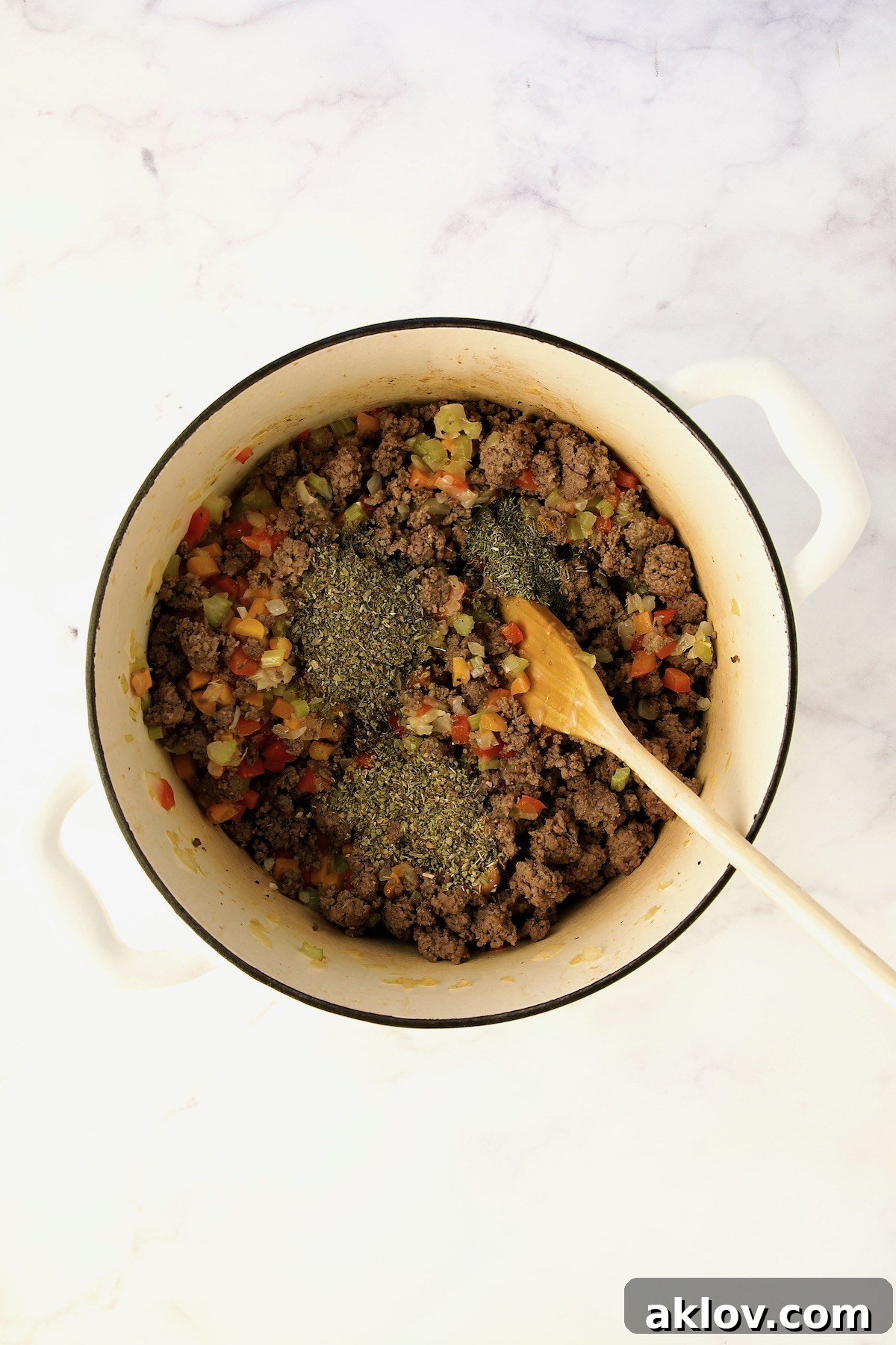 Dried herbs added to the mixture of ground beef and vegetables in a white Dutch oven.