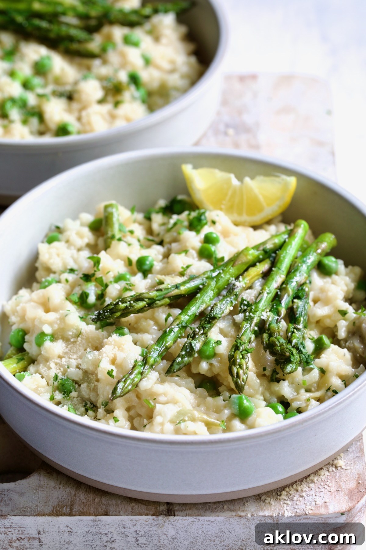 One bowl of Easy Risotto Recipe with Spring Vegetables with asparagus spears on the top and lemon wedge on whitewashed cutting board.