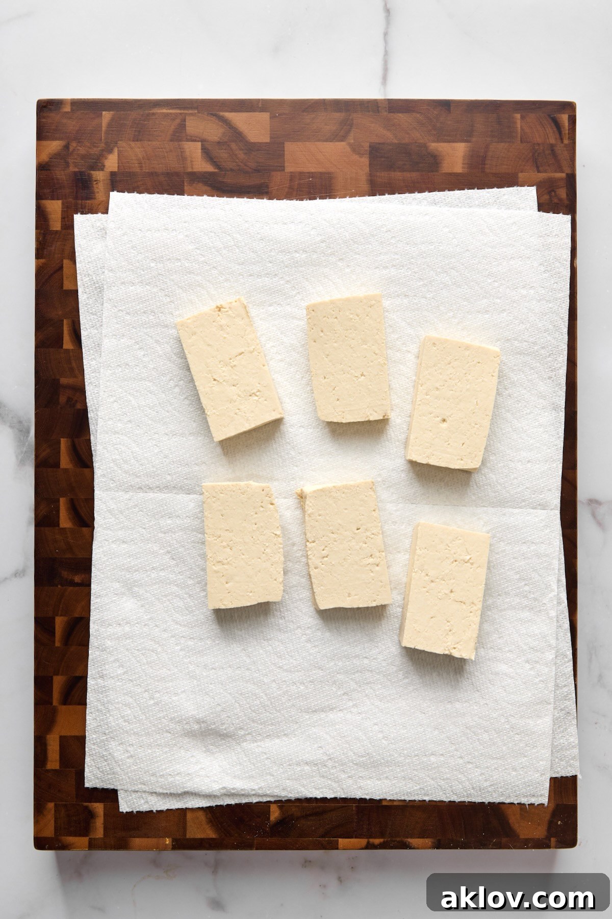 Six evenly cut slabs of extra-firm tofu resting on paper towels atop a wooden cutting board, prepared for pressing.