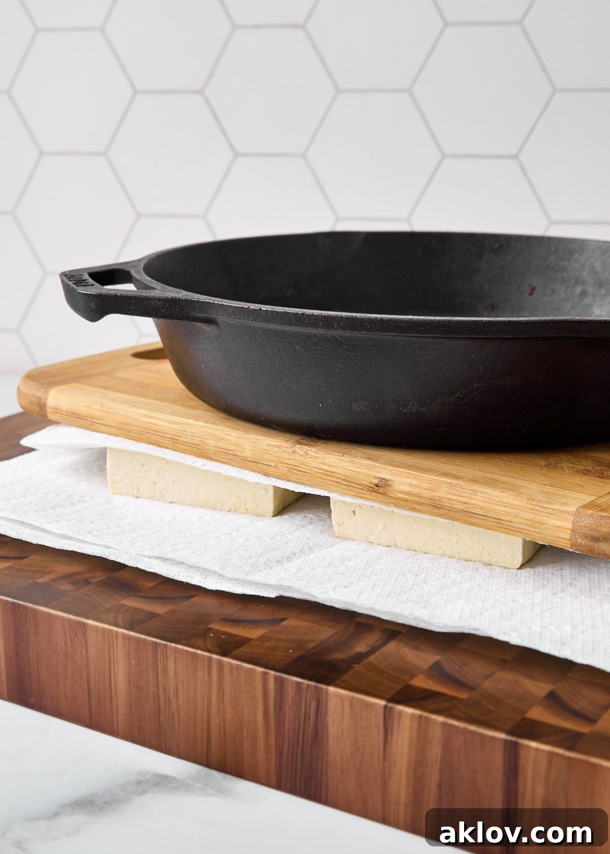 A heavy cast-iron skillet placed over layers of paper towels and tofu on a wooden cutting board, demonstrating the tofu pressing process.