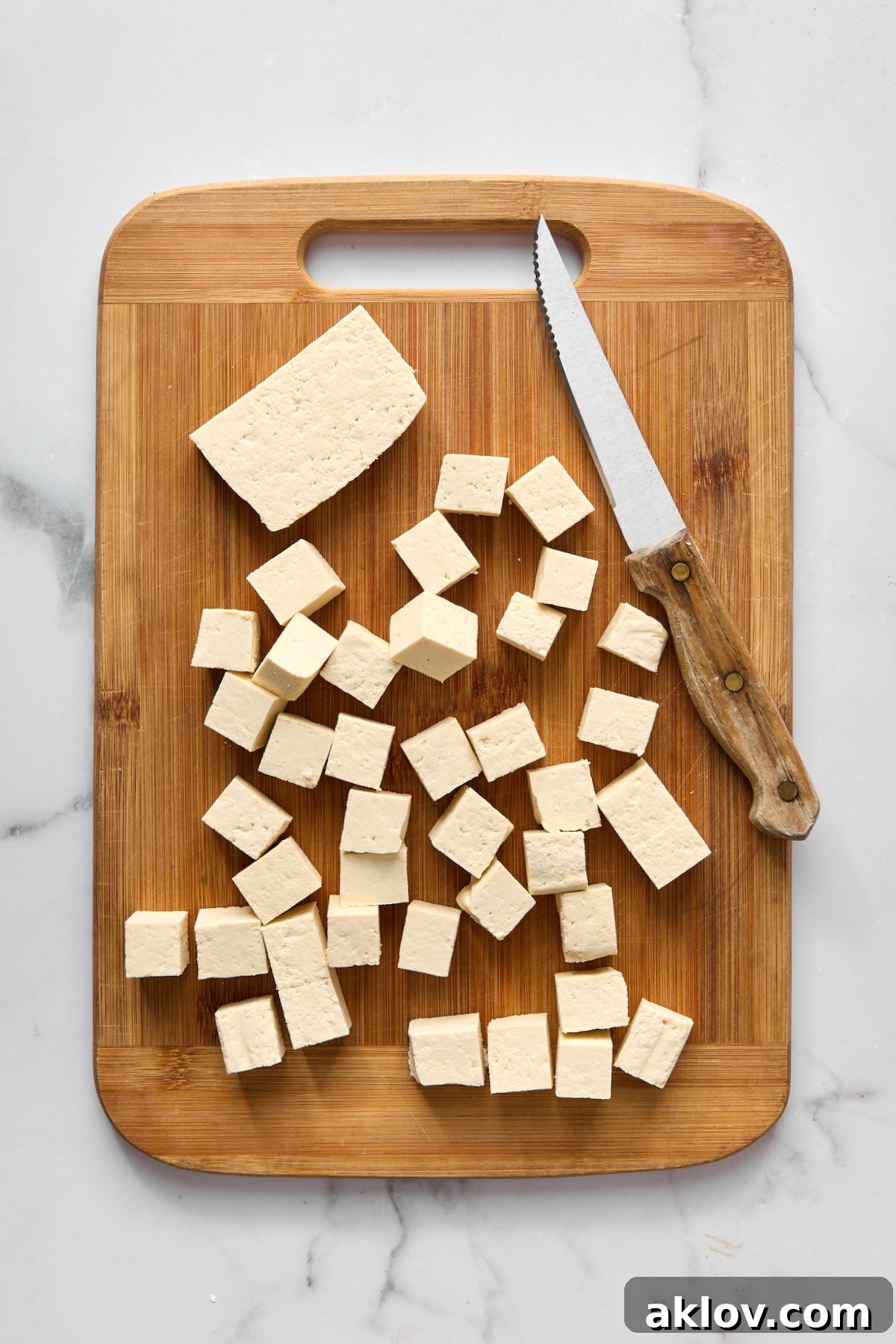 Pressed tofu cut into uniform 1/2-inch cubes with a knife on a bamboo cutting board, ready for seasoning.