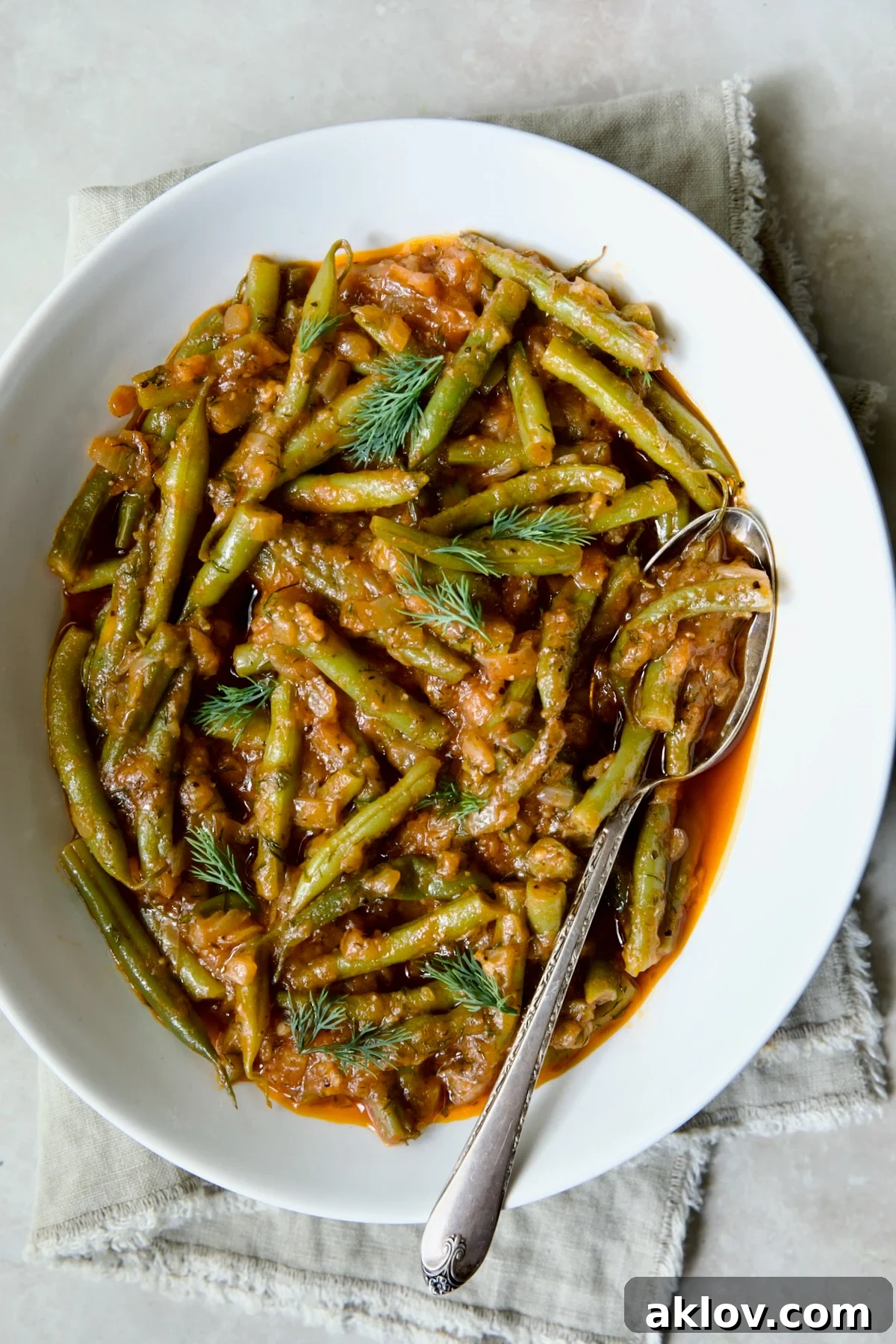 A serving of Greek Green Beans in a white oval bowl, garnished with fresh dill, resting on a beige frayed napkin.