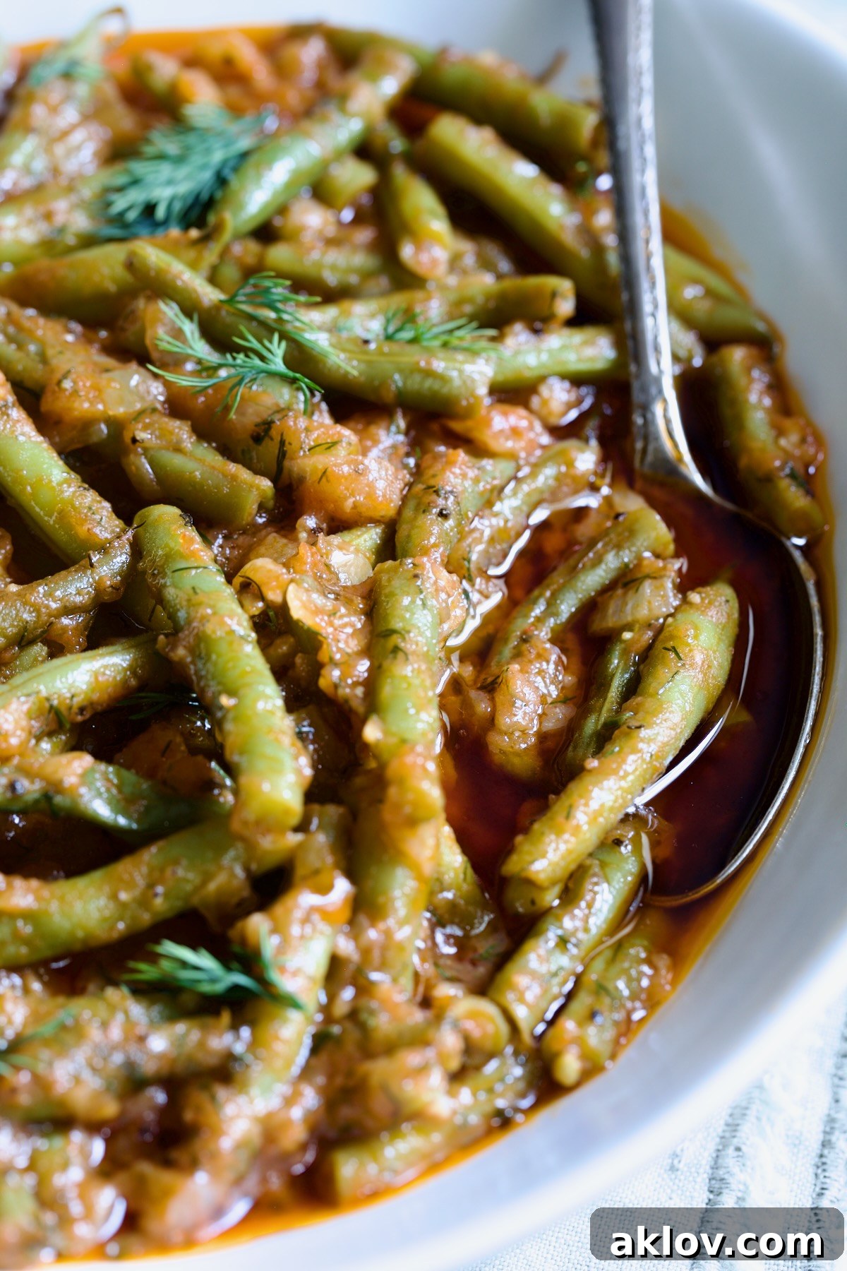 A close-up of Greek Green Beans in a white bowl with a serving spoon, highlighting their rich, tender appearance.