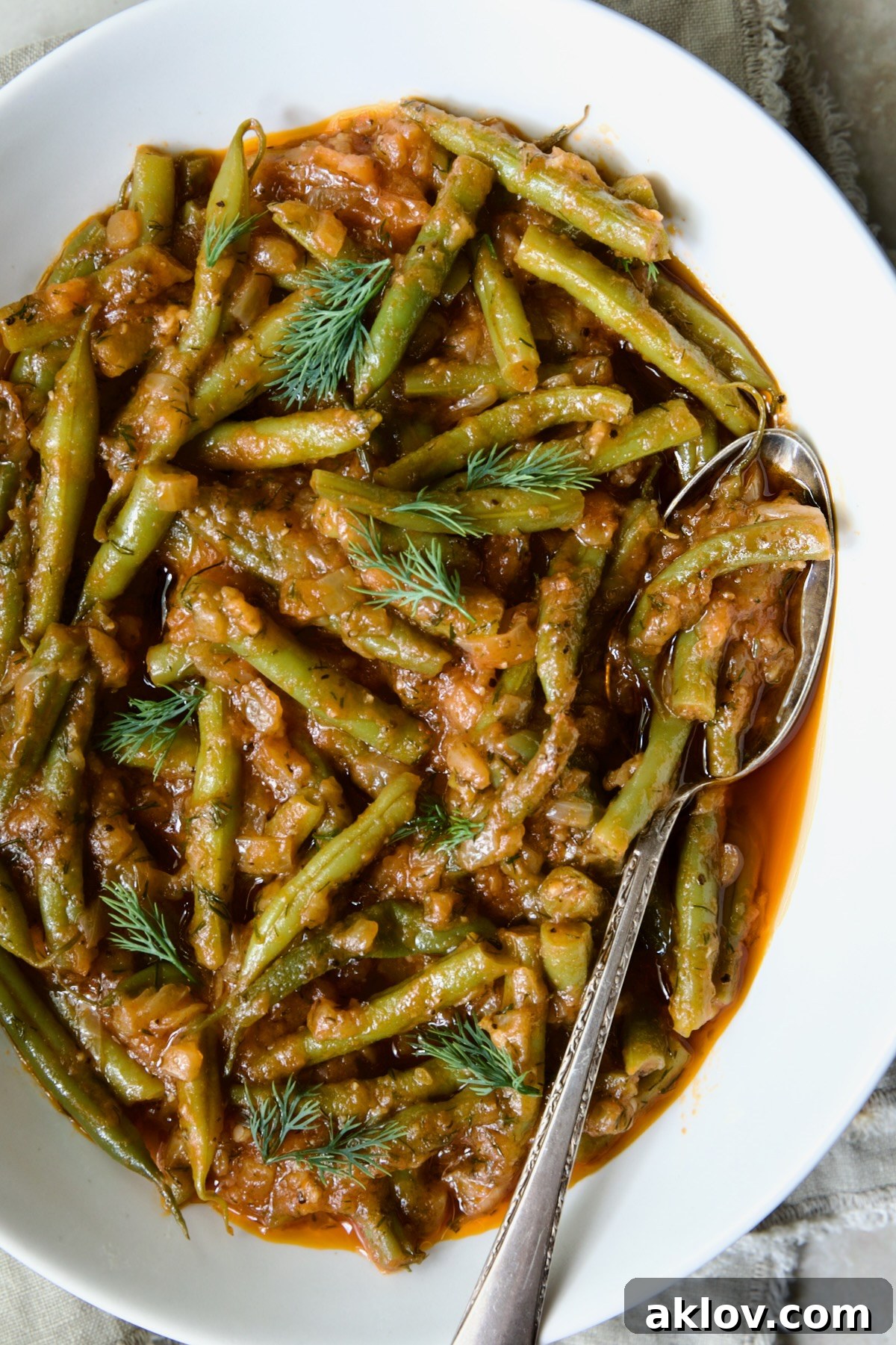 Greek Green Beans presented in a white serving bowl with a serving spoon, garnished with a sprig of fresh dill.