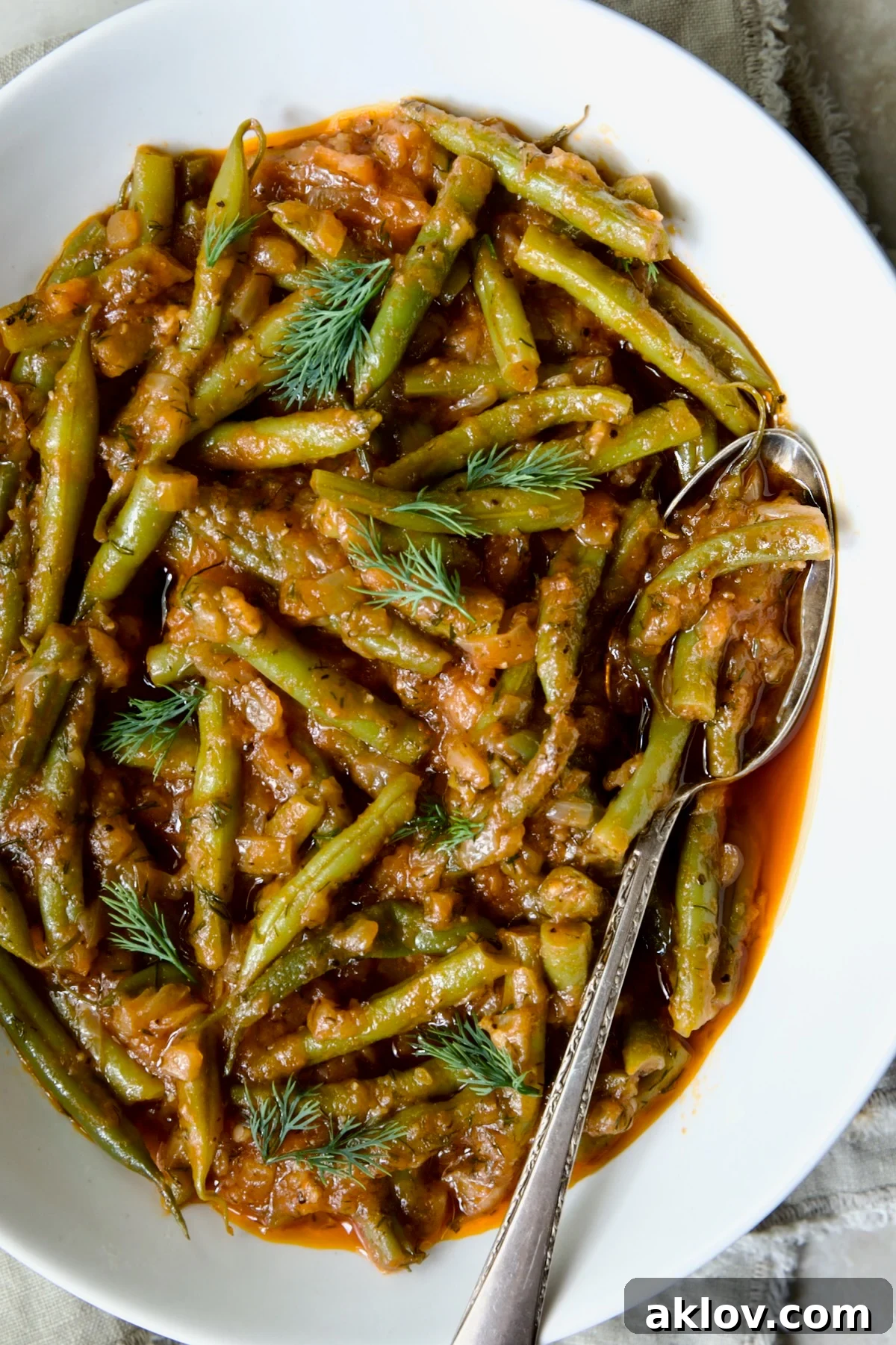 Greek Green Beans in a white serving bowl with a serving spoon, garnished with fresh dill sprigs.