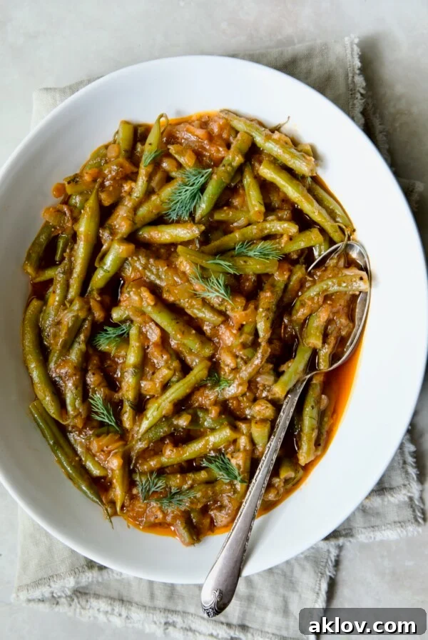 Greek Green Beans in white oval serving bowl with serving spoon on beige frayed napkin.