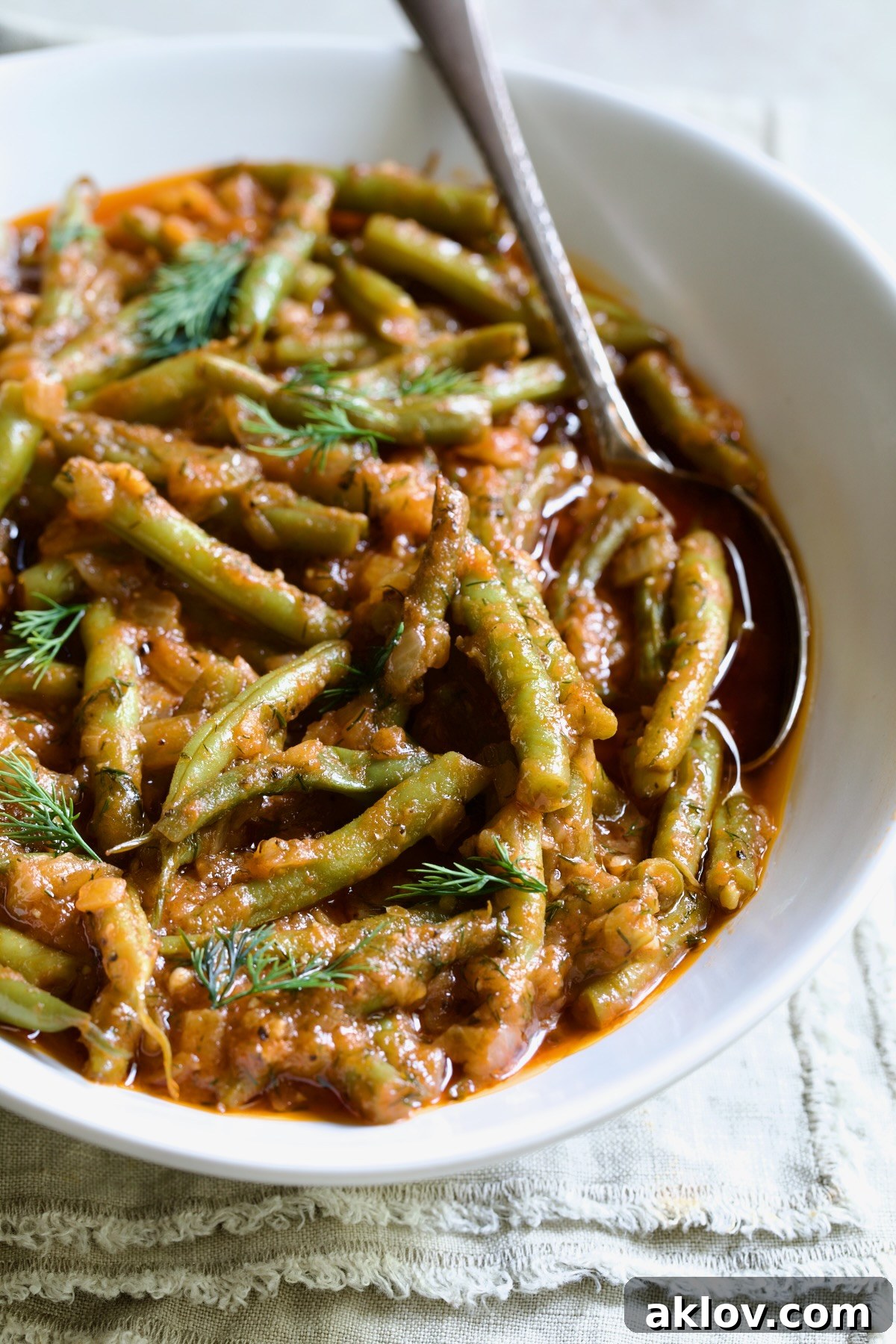 Greek Green Beans in a white serving bowl, garnished with fresh dill, accompanied by a serving spoon on a frayed napkin.
