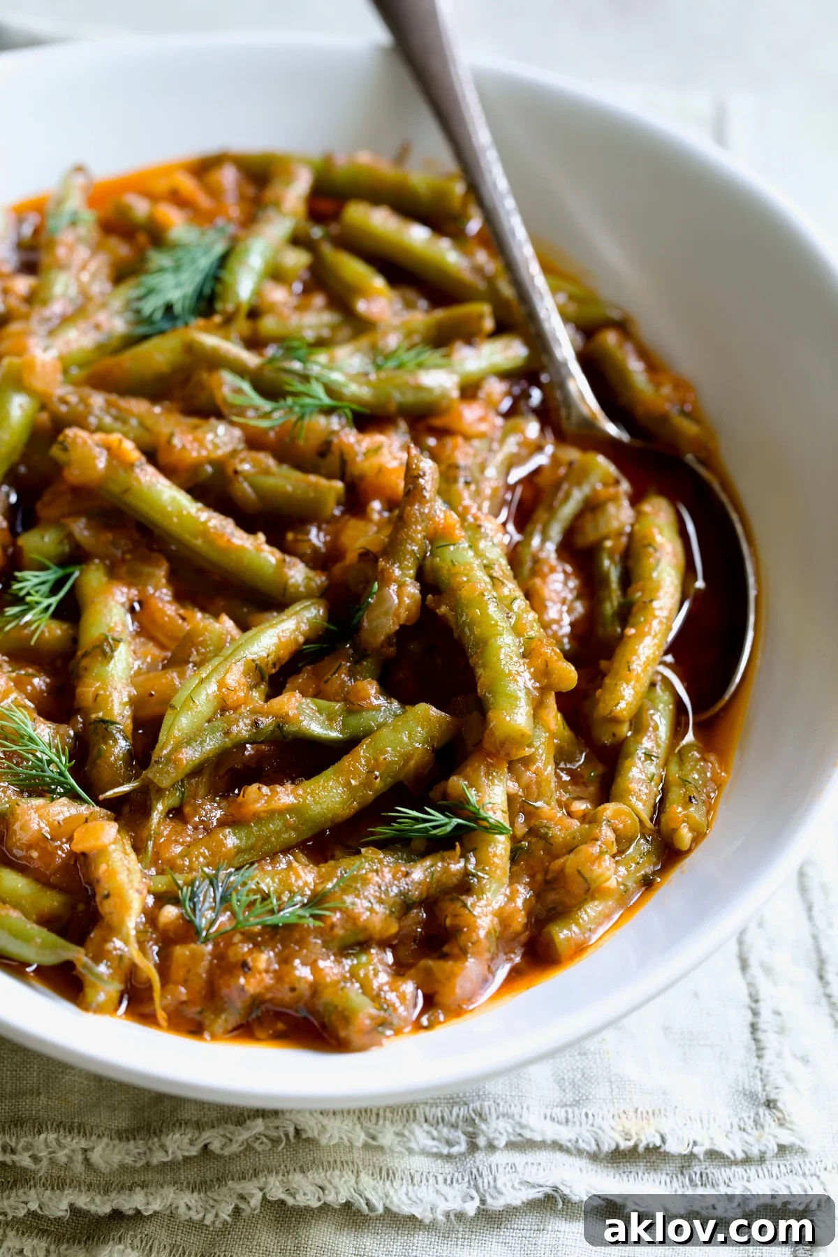 Greek Green Beans in white serving bowl with serving spoon garnished with fresh dill on frayed napkin, close-up.