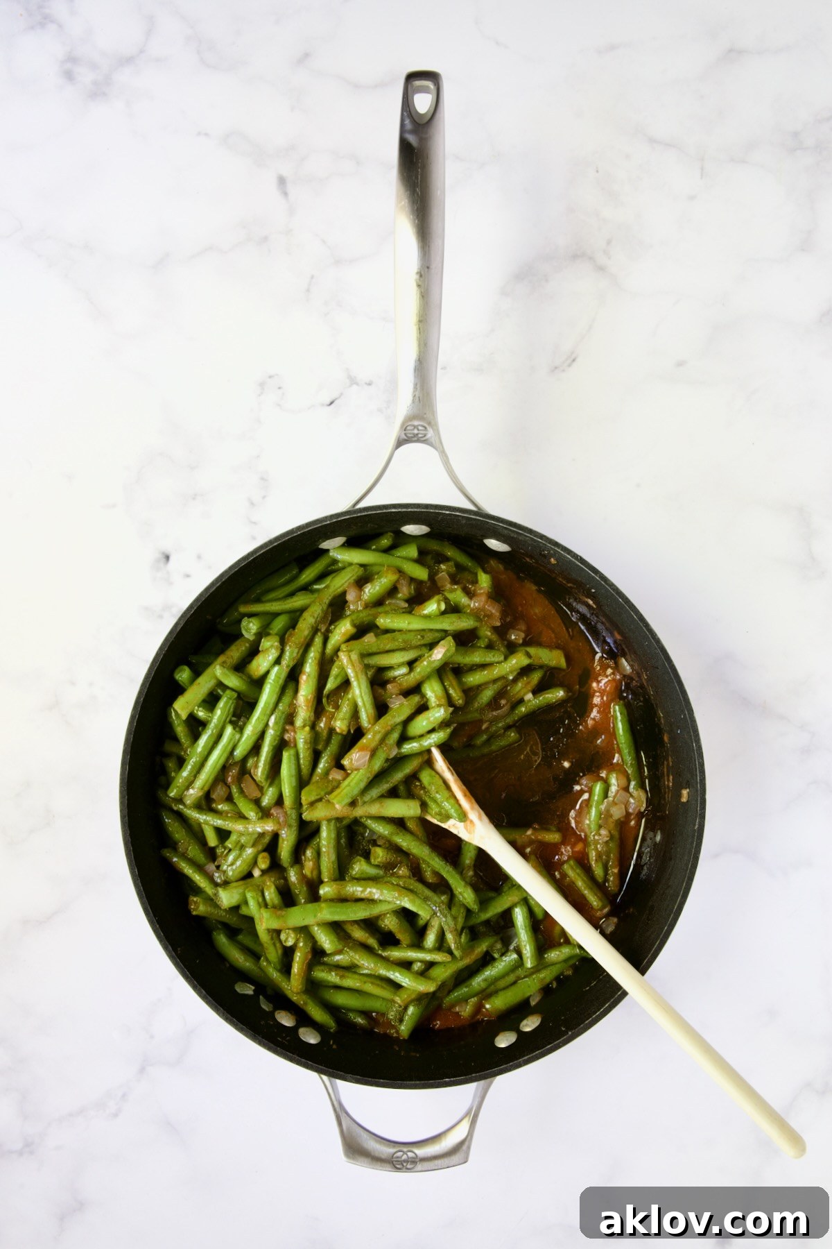 A white spoon stirring green beans, tomato sauce, and dill in a nonstick skillet, ensuring even cooking.