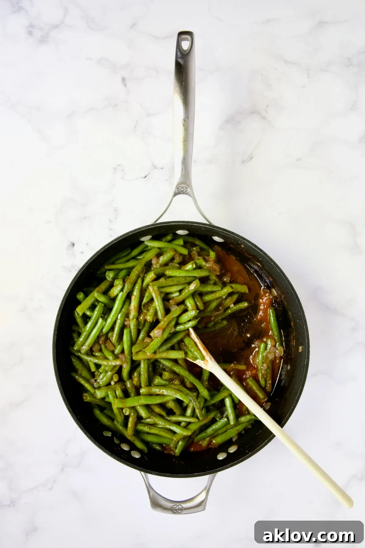 A white spoon stirring green beans, tomato sauce, and dill in a nonstick skillet.