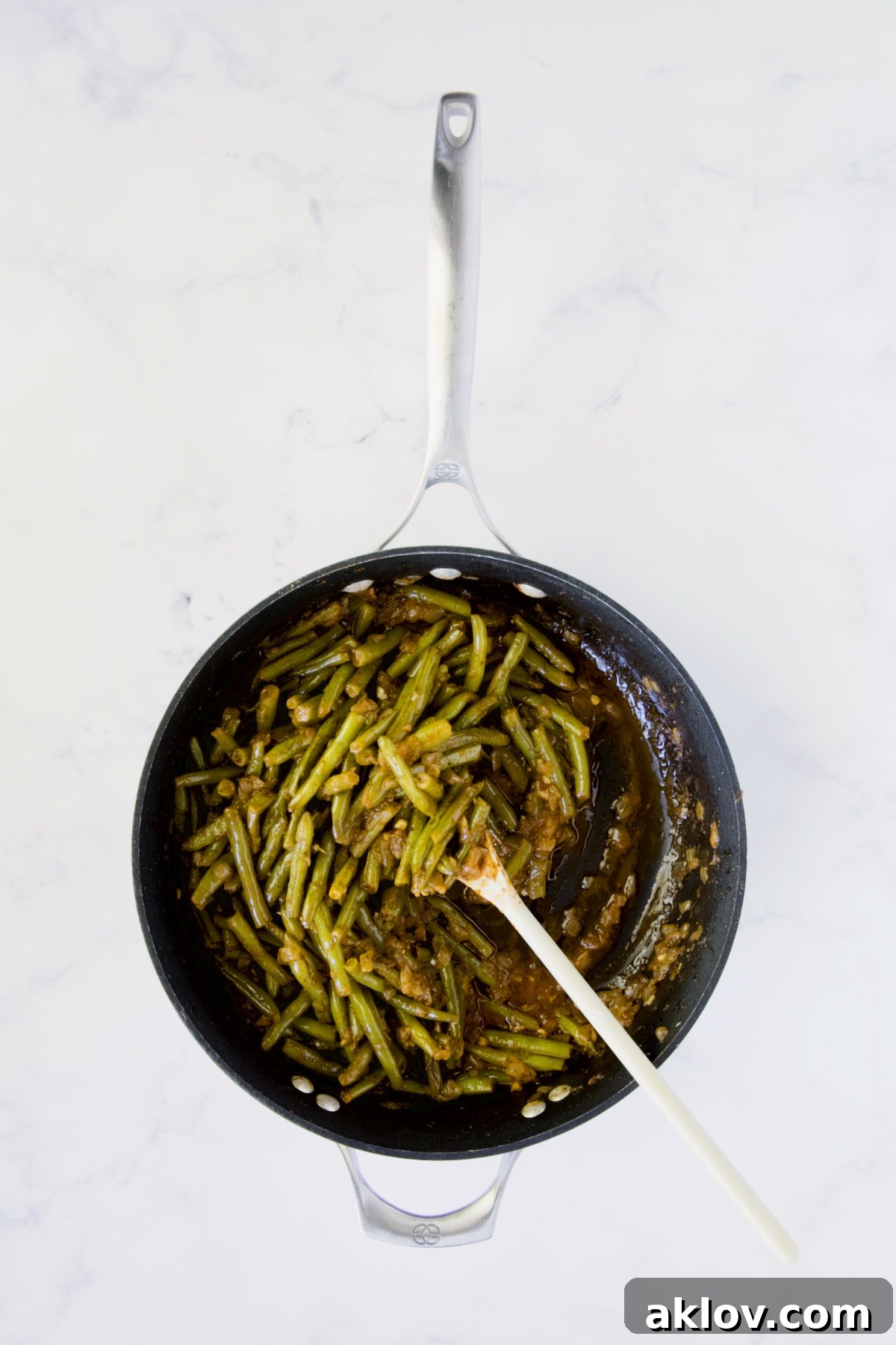 Finished Greek Green Beans in a nonstick skillet, being gently stirred with a white spoon.