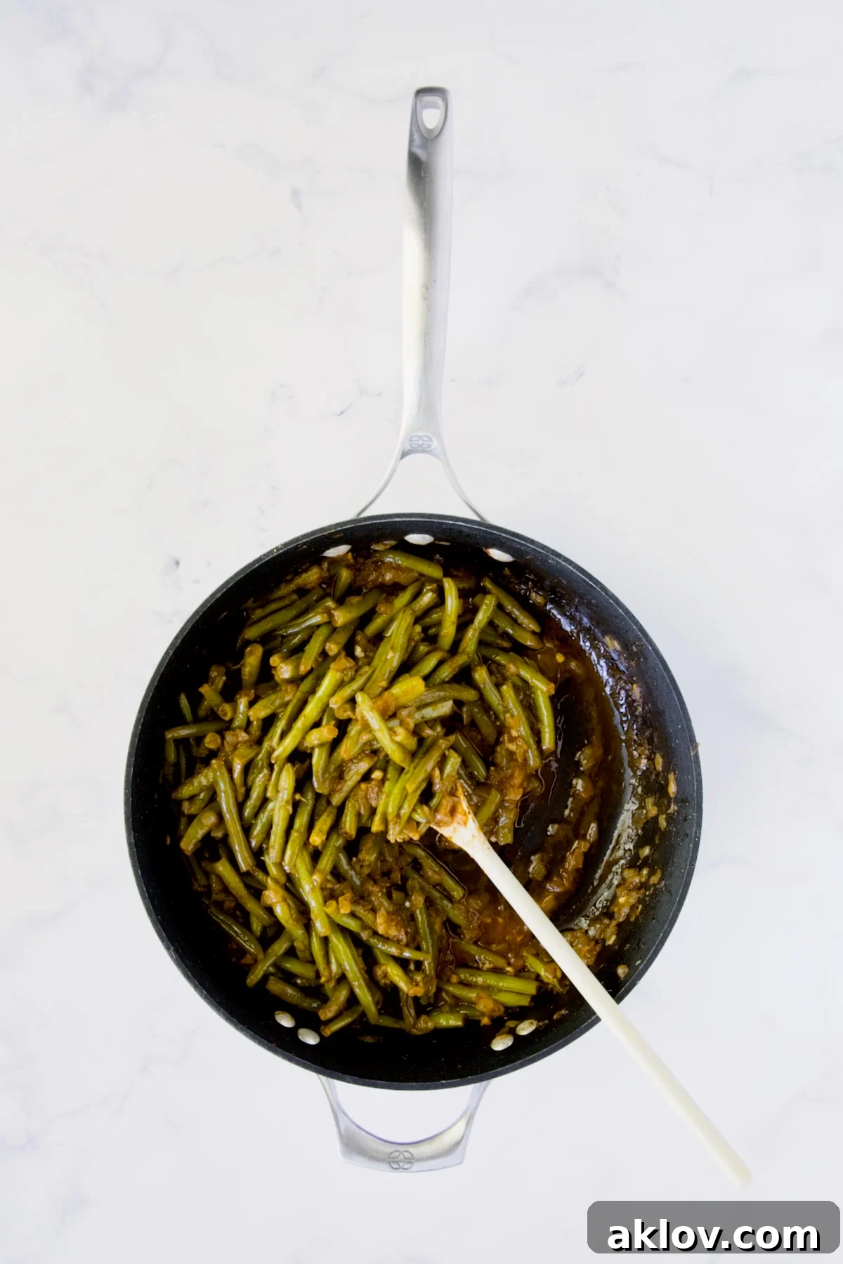 Finished Greek Green Beans in a nonstick skillet being stirred with a white spoon.