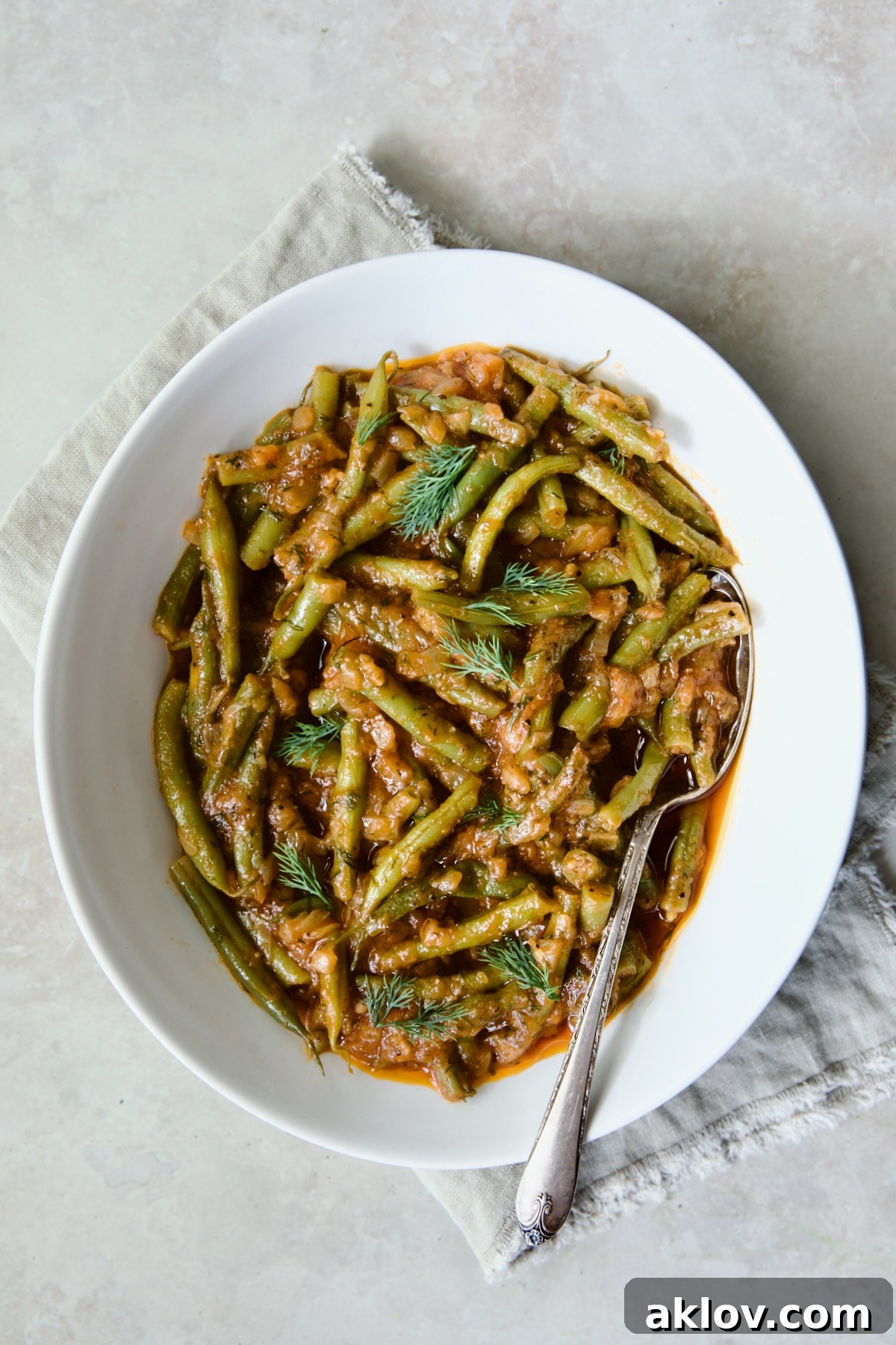 A close-up of Greek Green Beans in a white oval serving bowl with a serving spoon on a frayed napkin, showcasing their tender texture.