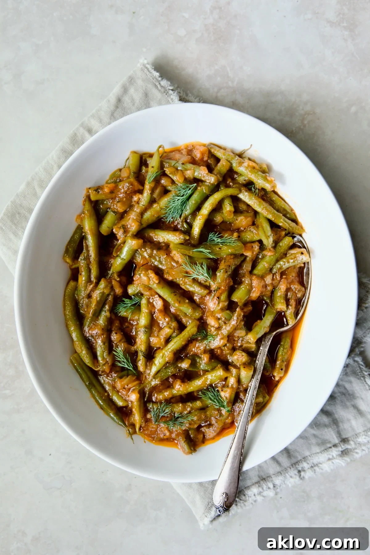 Greek Green Beans in a white oval serving bowl with a serving spoon on a frayed napkin.