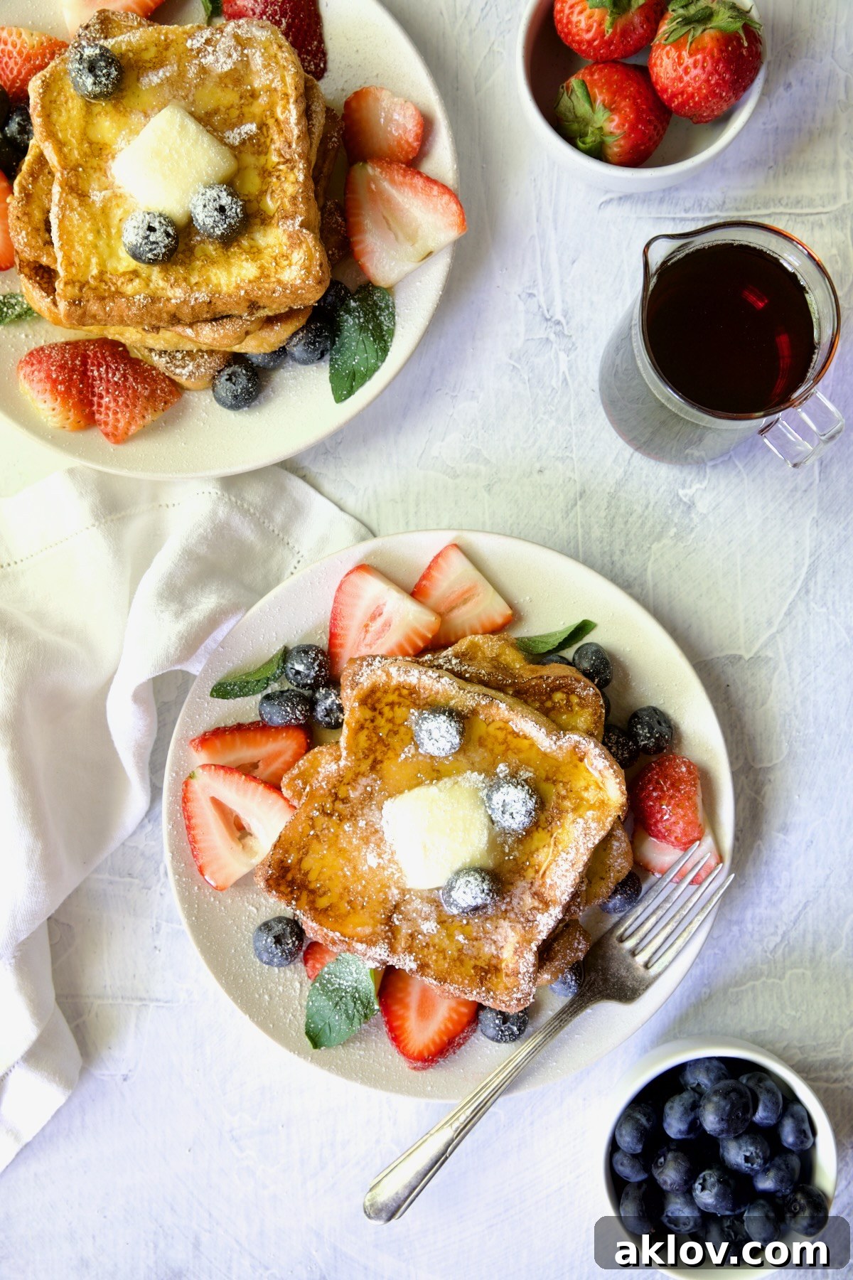 Two plates of beautifully garnished Brioche French Toast with fresh fruit, a pat of butter, and powdered sugar.