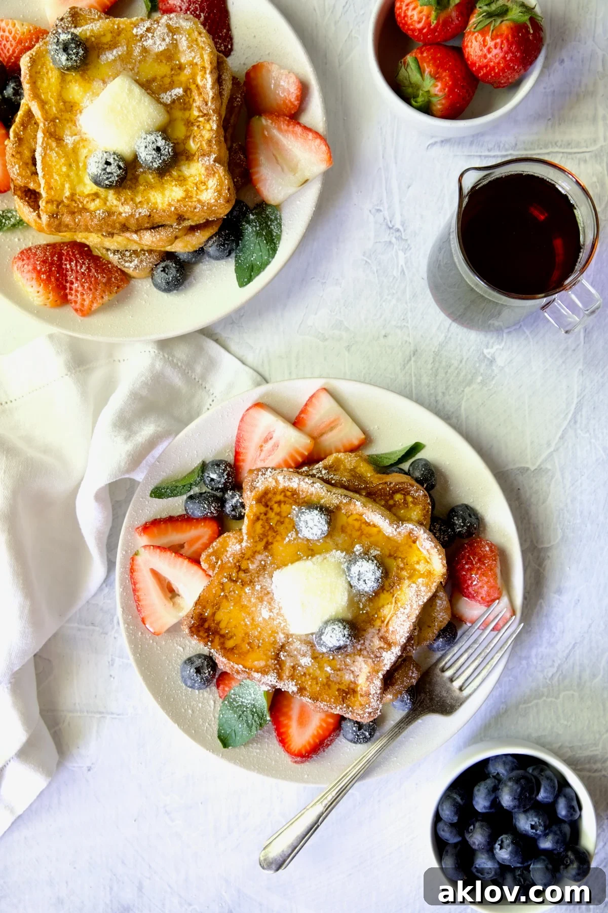 Two plates of Brioche French Toast garnished with fresh fruit, a pat of butter, and powdered sugar, identical to the previous image but serving as a visual break.