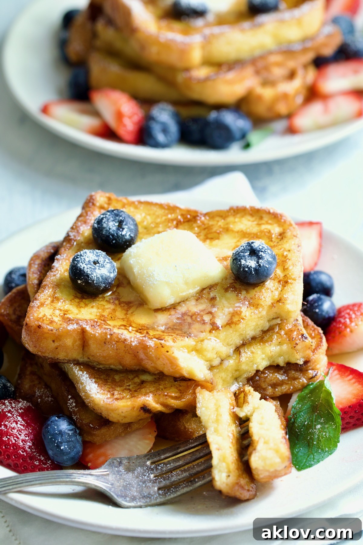 Brioche French Toast on white plate with pat of butter, blueberries and strawberries. Two pieces are on a fork, showing the fluffy interior.