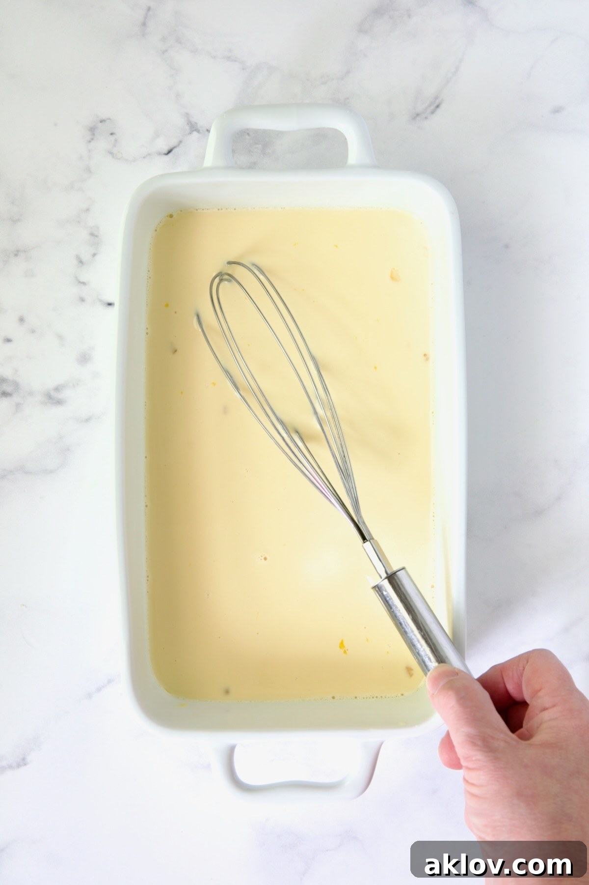 Rich vanilla custard being whisked in a white rectangular dish with a wire whisk.