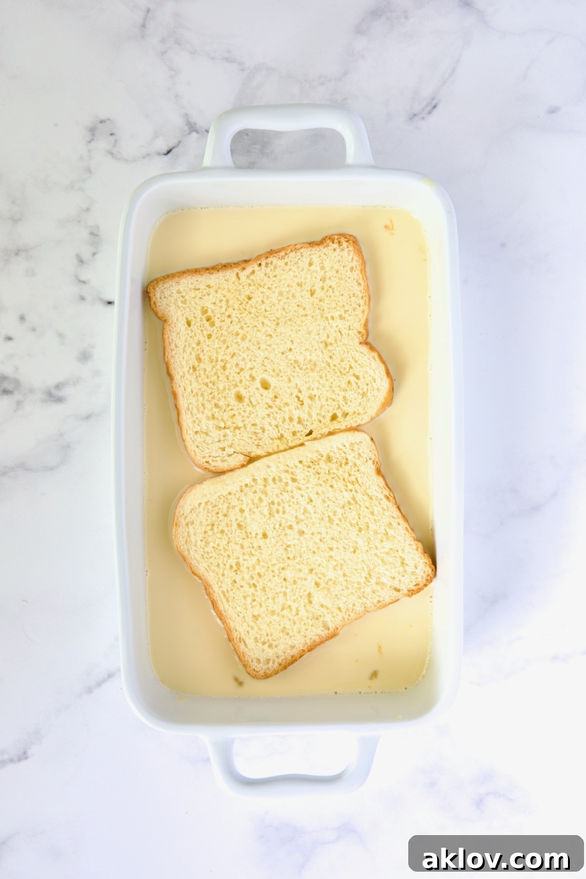 Two slices of brioche bread soaking in the vanilla custard mixture in a white rectangular baking dish.