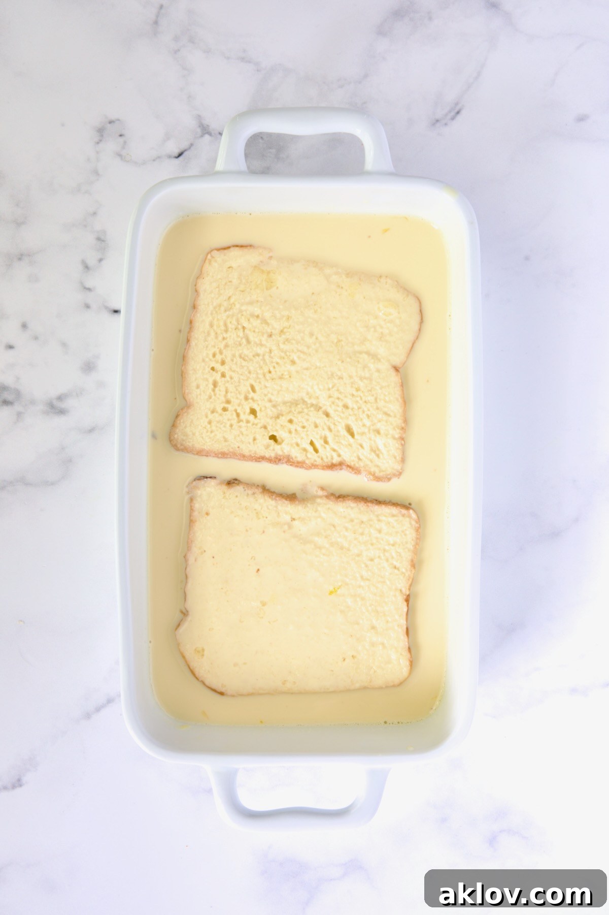 Two slices of brioche bread after being flipped, fully saturated in the custard, in a white rectangular baking dish.