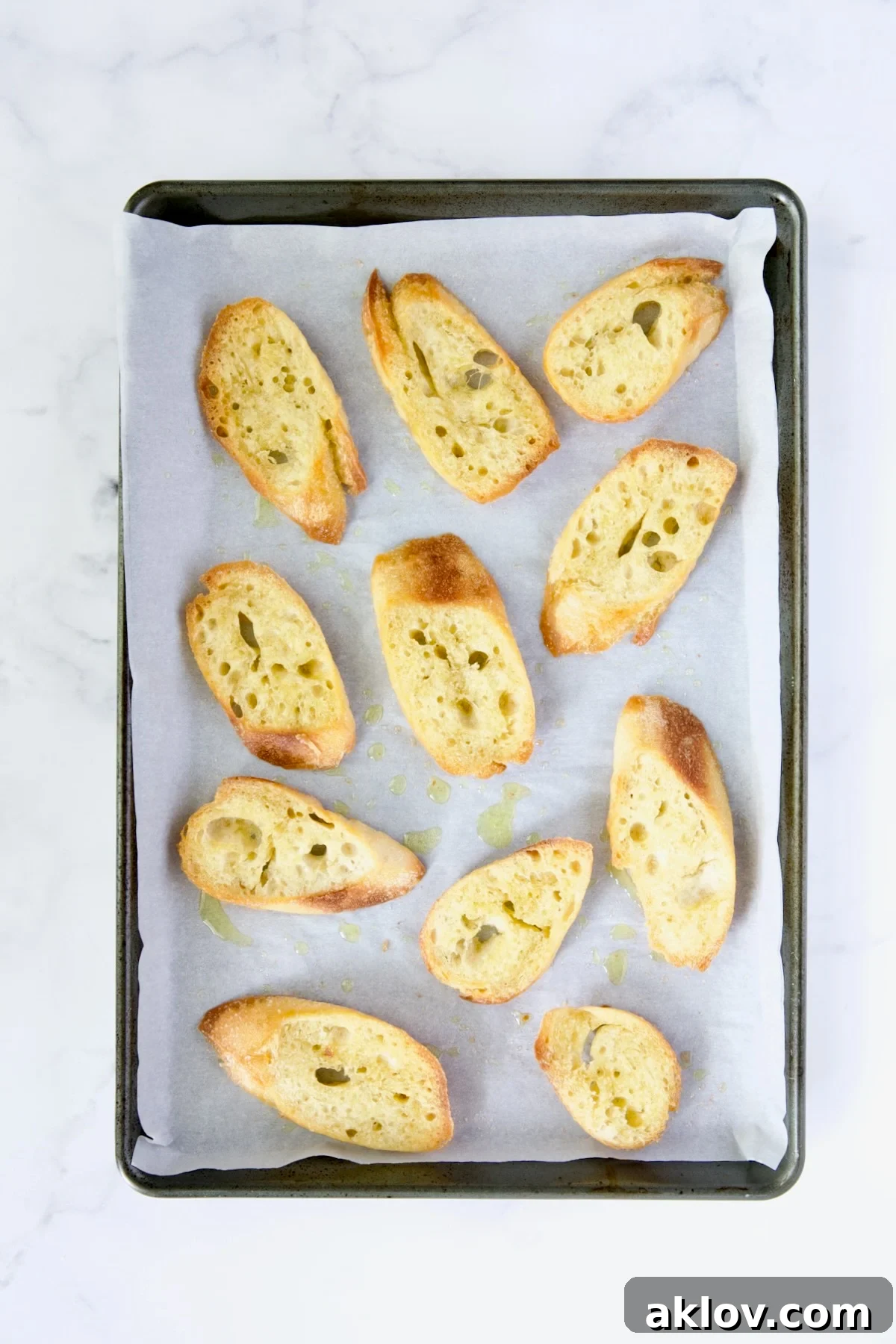 Eleven pieces of baguette slices brushed with olive oil and lightly salted, arranged on parchment paper on a baking sheet.
