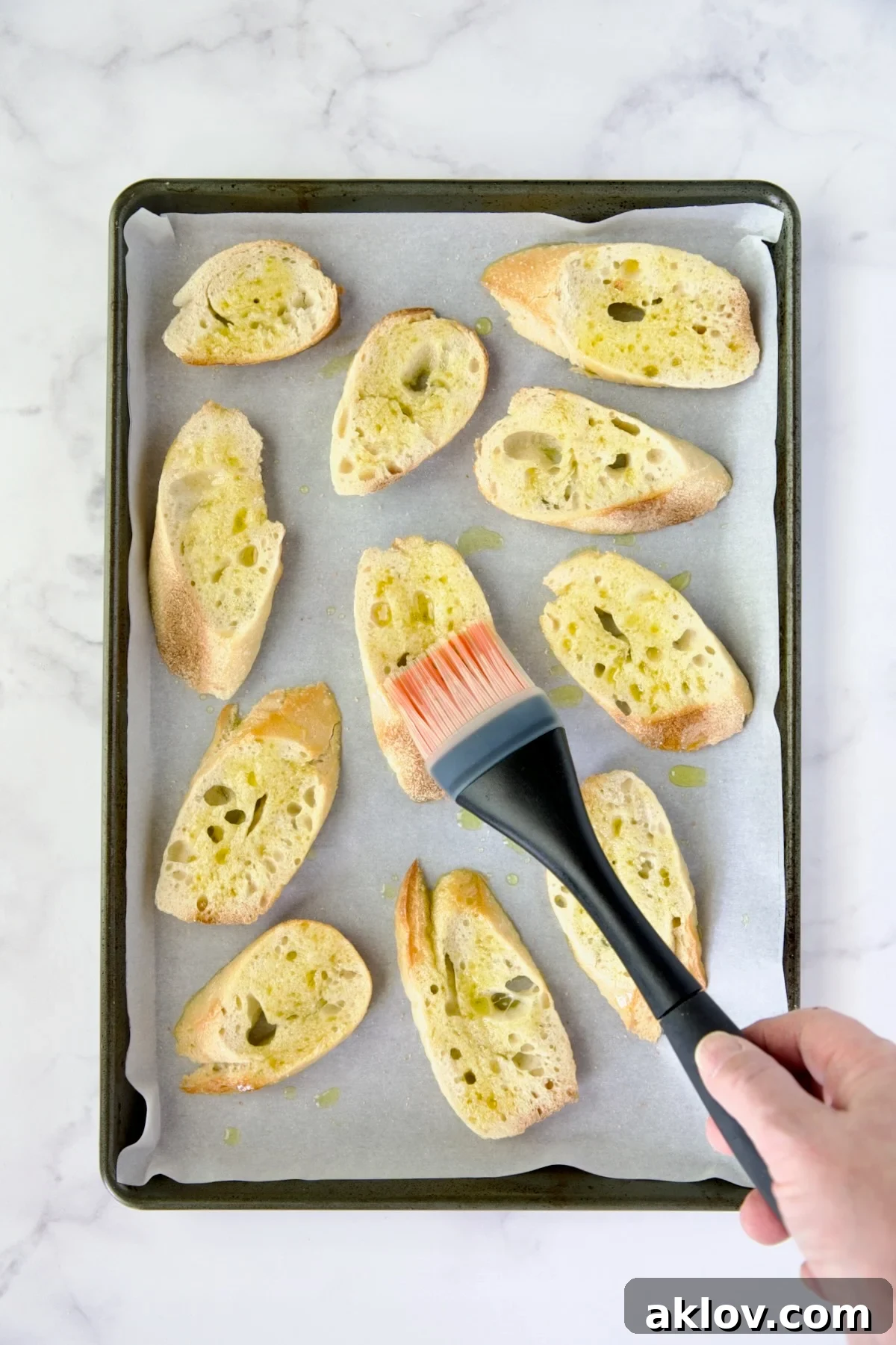 Hand brushing olive oil onto baguette slices on a baking sheet, illustrating crostini preparation.