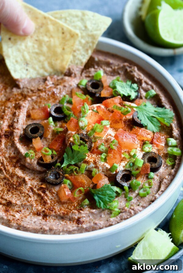 Black Bean Dip garnished with black olives, tomatoes, scallions and cilantro leaves in white clay bowl with tortilla chips and lime wedges.