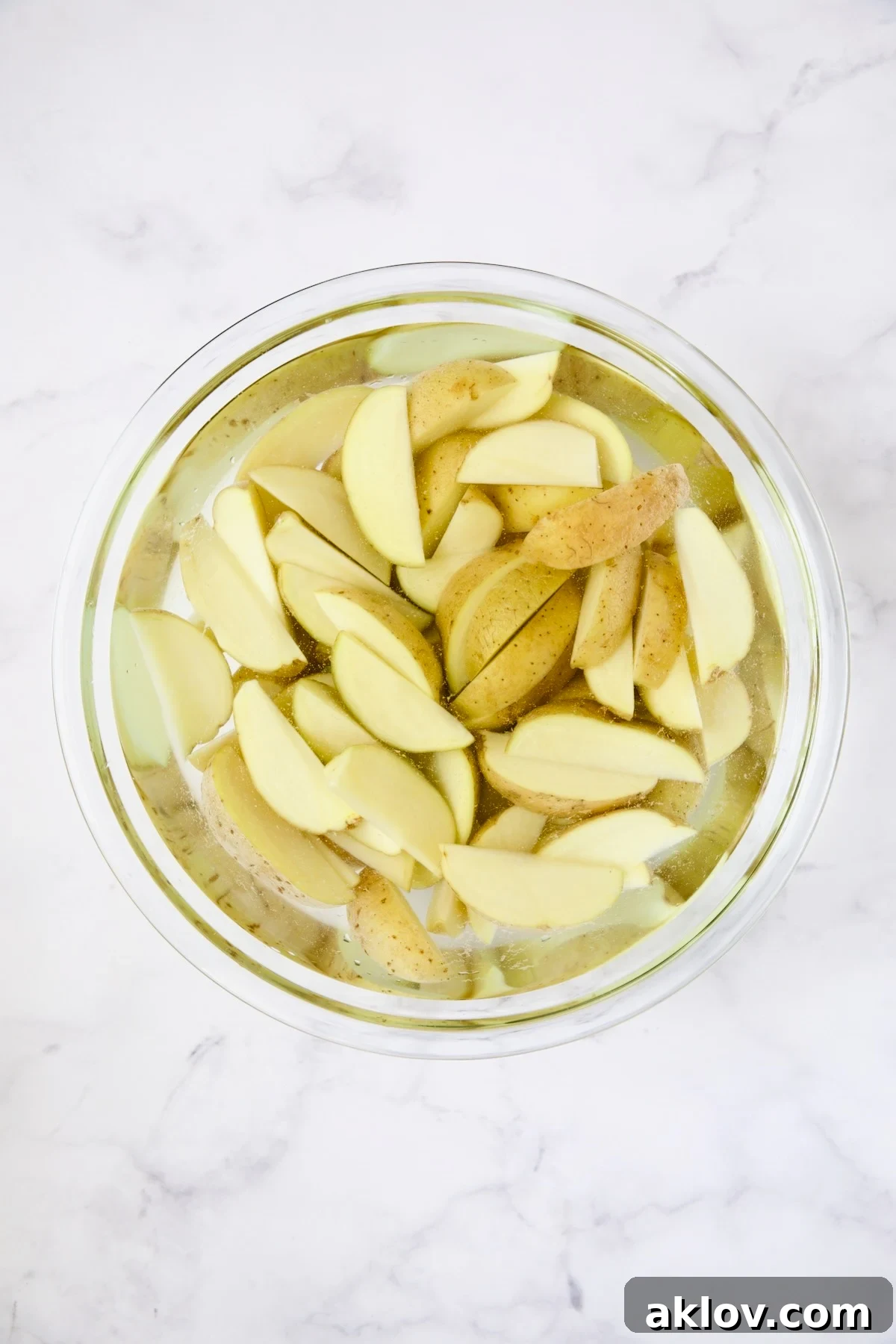Freshly cut Yukon Gold potato wedges soaking in cold water in a clear glass bowl, a technique for extra crispiness.