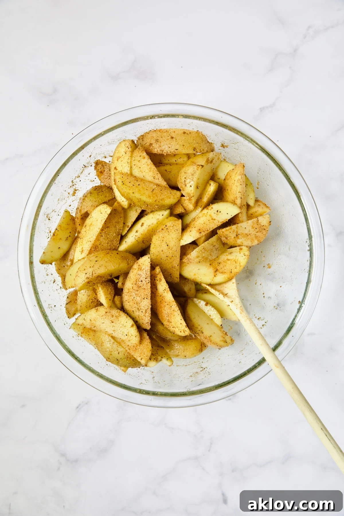 Seasoned Yukon Gold potato wedges being stirred with a white spoon in a glass bowl, showcasing thorough spice distribution.
