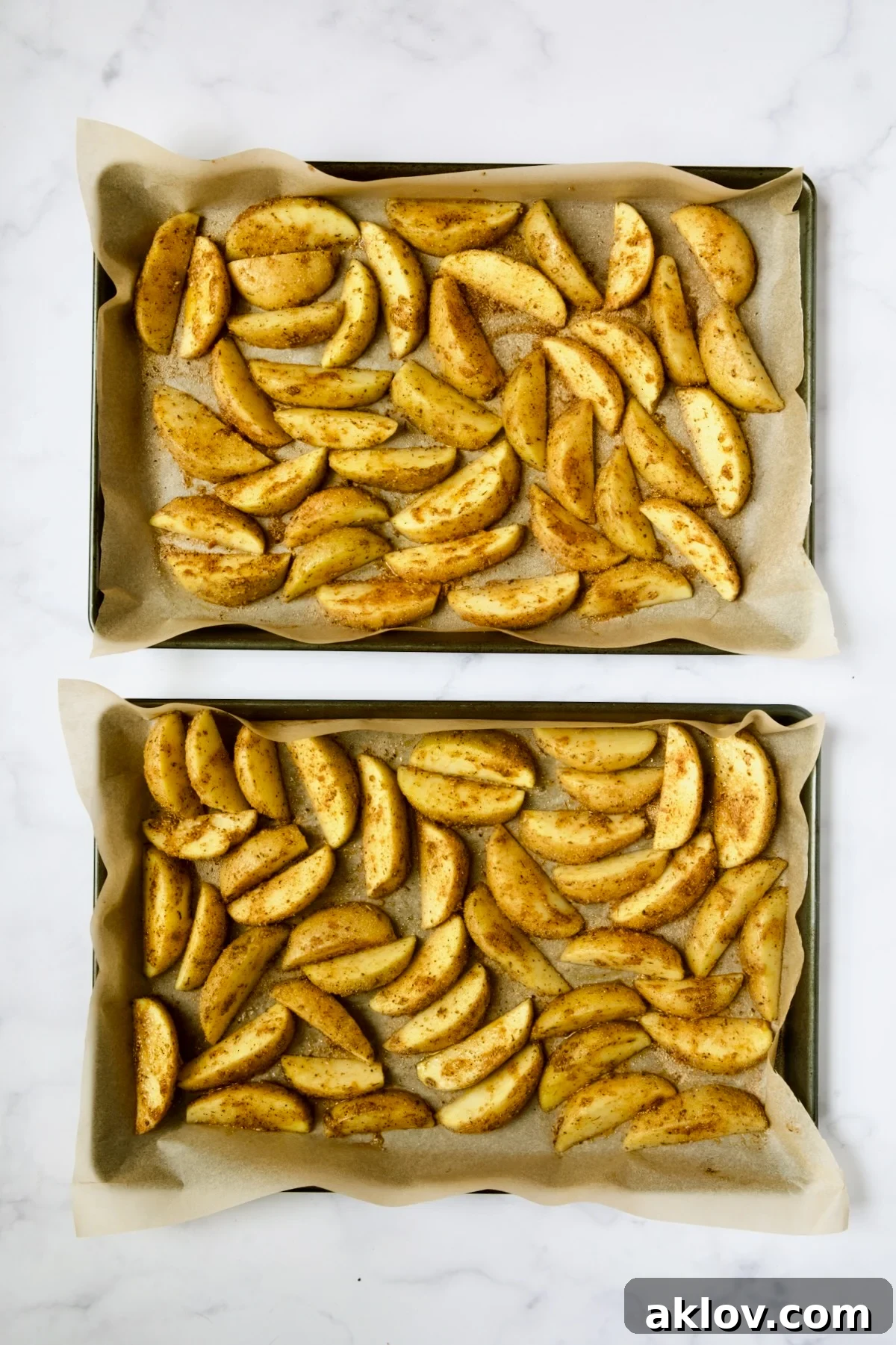 Two baking sheets meticulously lined with parchment paper, holding evenly spaced seasoned potato wedges, ready for roasting.