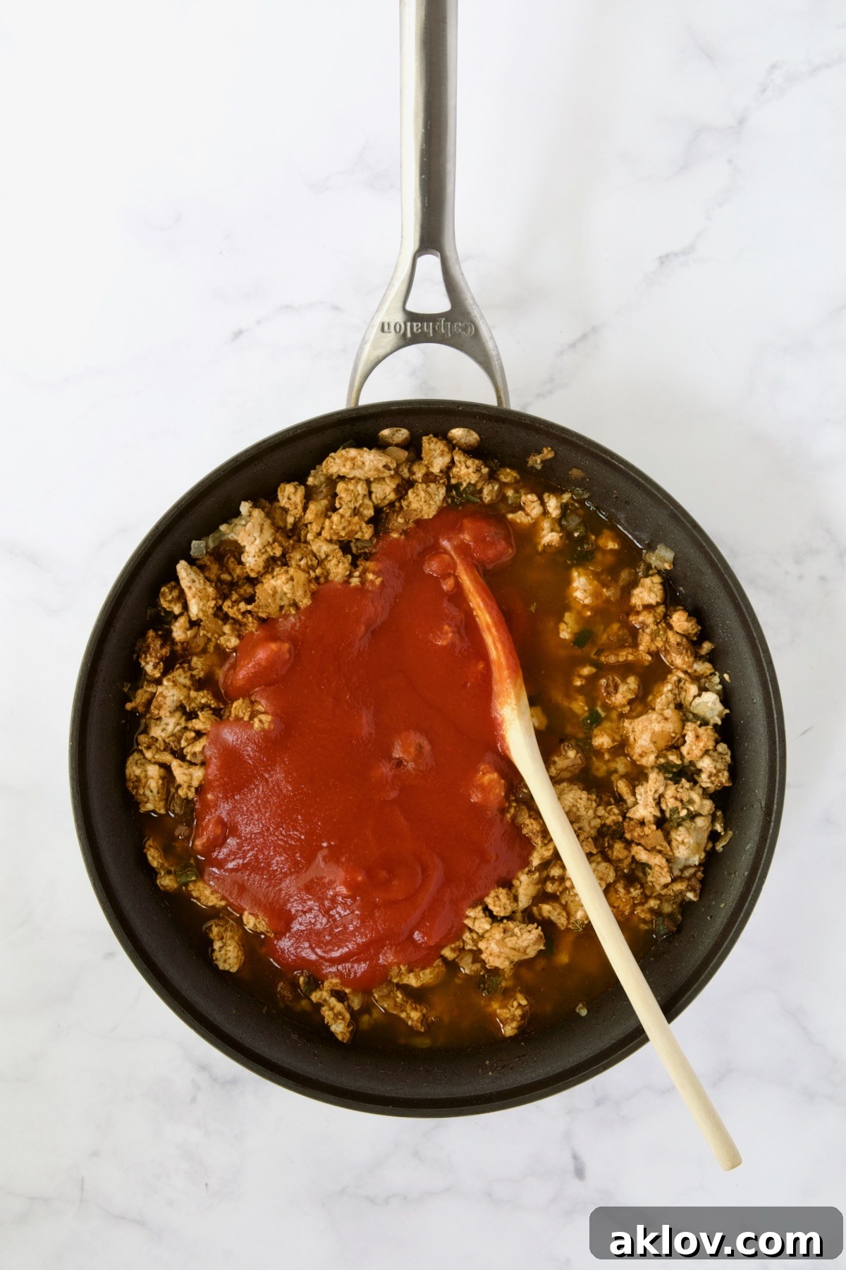 Tomato sauce added to ground chicken in nonstick skillet being stirred with wooden spoon.