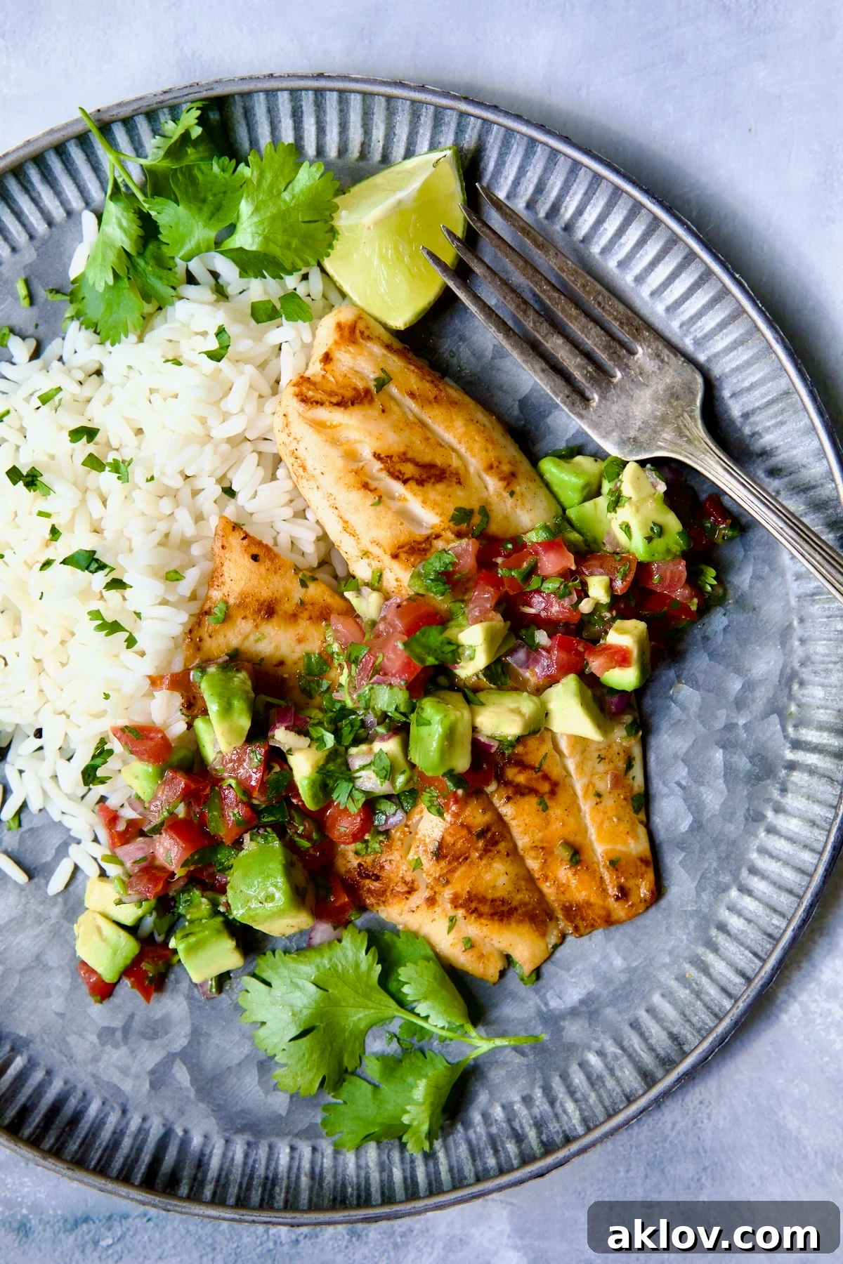 Grilled Tilapia with Tomato - Avocado Salsa on a round metal plate with white rice, garnished with cilantro and a lime wedge.