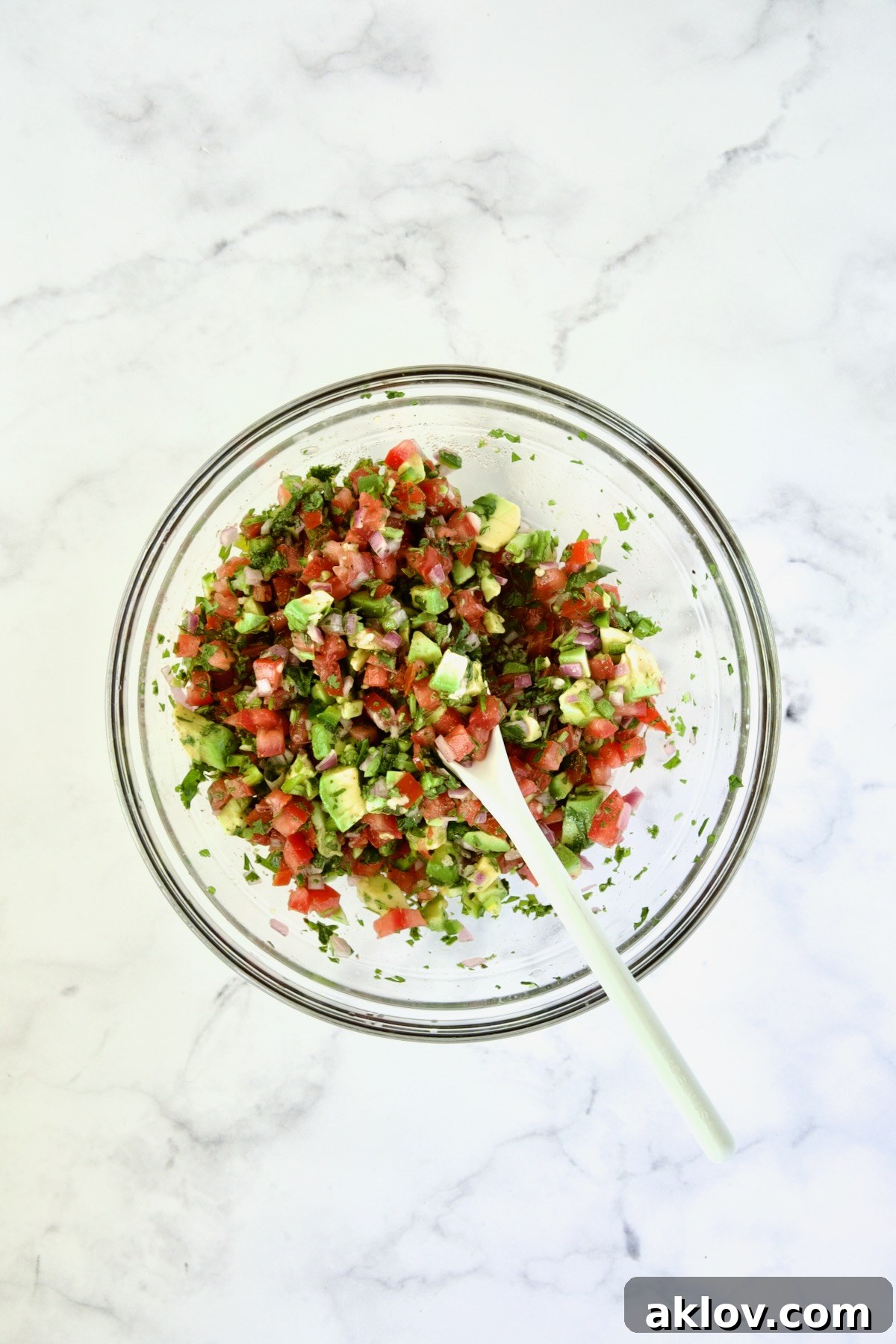 Tomato - Avocado Salsa in glass bowl being stirred with a white spoon.