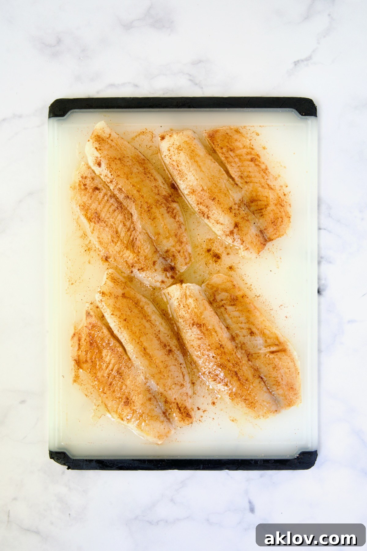 Four pieces of seasoned tilapia fillets on a white plastic cutting board, ready for grilling.