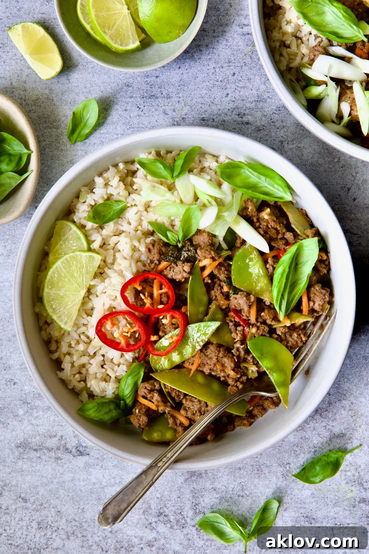 A beautifully plated bowl of Thai Basil Beef stir-fry, garnished with fresh lime wedges, vibrant basil leaves, and thinly sliced Fresno chiles. The rich, glossy sauce coats the ground beef and crisp vegetables, hinting at the dish's incredible flavor.