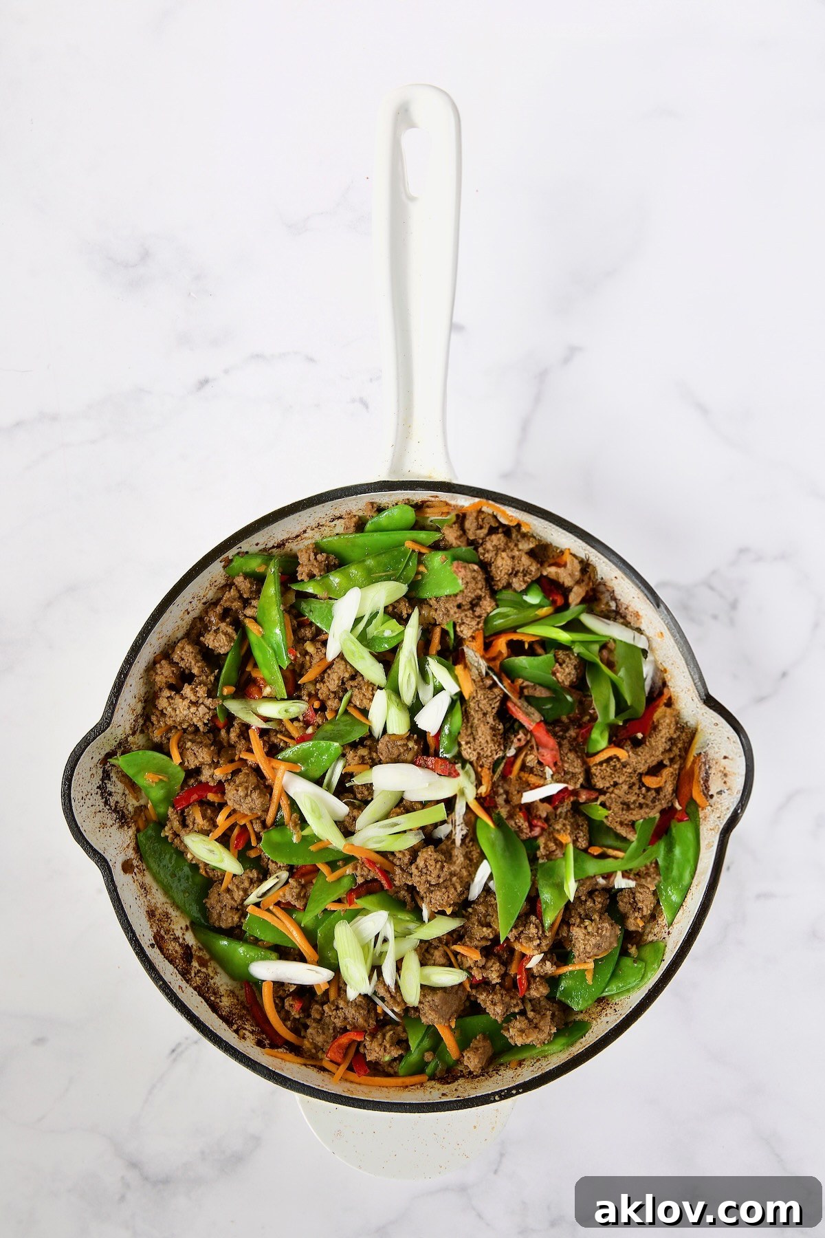 The stir-fry in a white enamel cast iron skillet, now with scallions added, combining with the ground beef, Fresno chiles, shredded carrots, and snow peas for a burst of color and flavor.