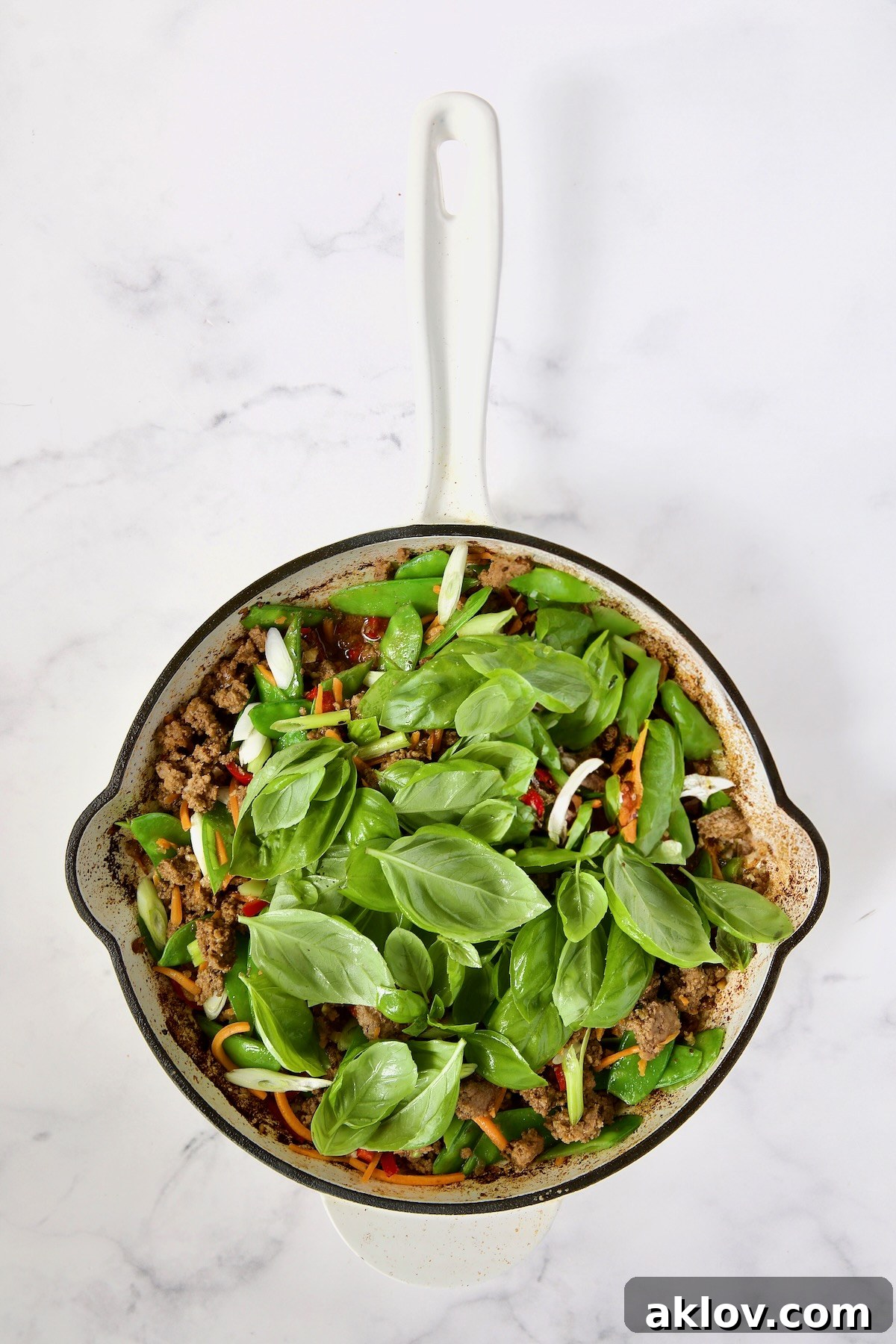 The completed Thai Basil Beef stir-fry, with fresh basil leaves now incorporated, creating a fragrant and visually appealing dish in the white enamel cast iron skillet.