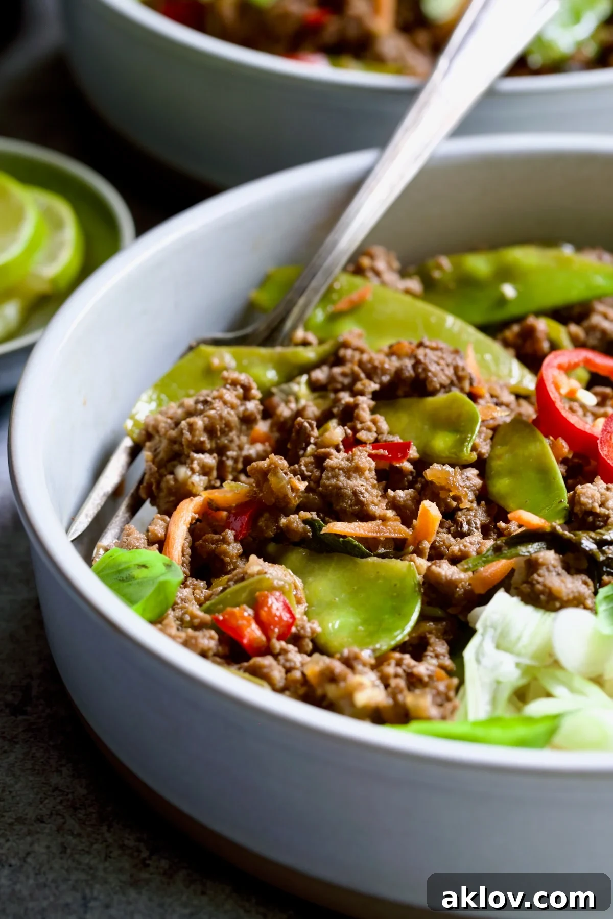 A close-up of Thai Basil Beef in a white bowl, showing the texture of the ground beef, the colorful vegetables, and the fragrant basil. A fork is poised, ready for a bite, emphasizing the dish's appetizing appeal.