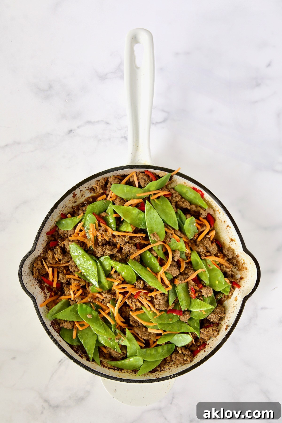 A colorful stir-fry in a white enamel cast iron skillet featuring cooked ground beef, Fresno chiles, shredded carrots, and snow peas, coated in a glistening sauce.