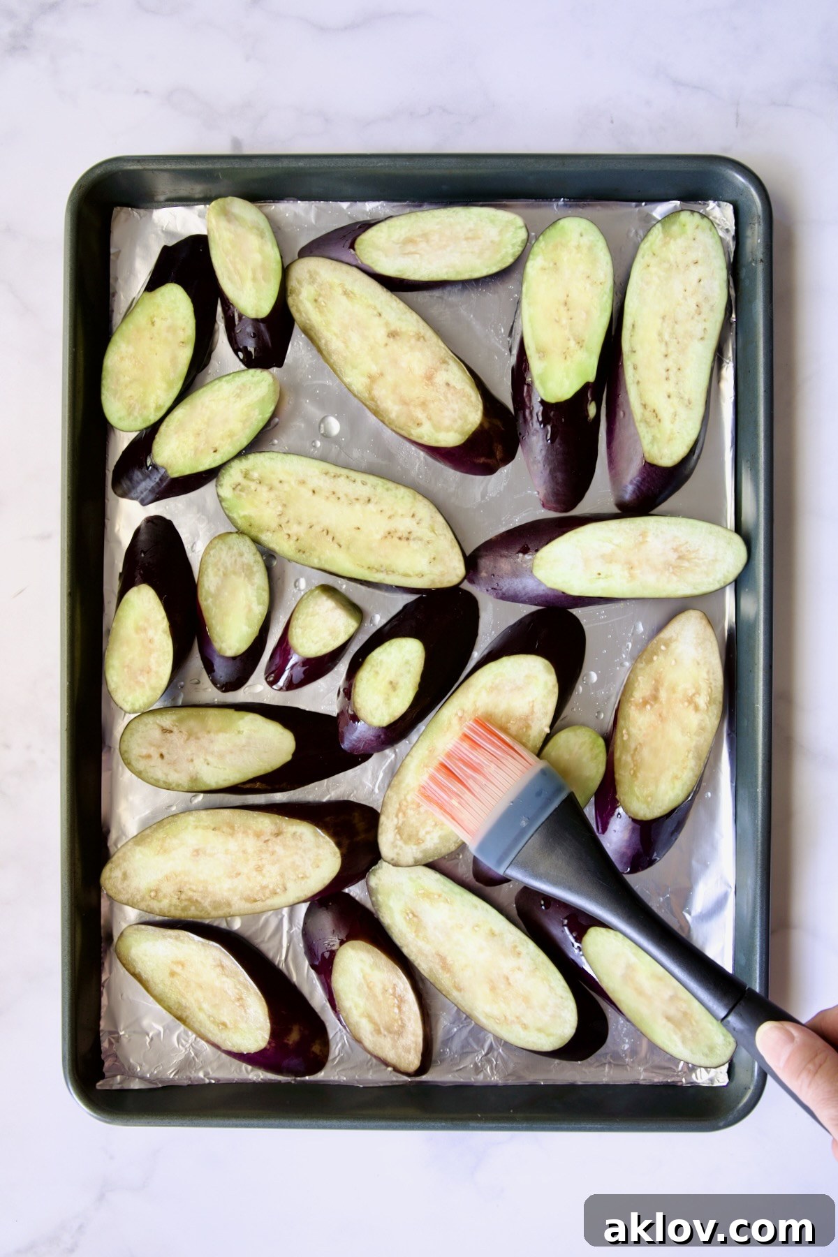 Slices of eggplant cut on the diagonal on aluminum foil-lined rimmed sheet pan being brushed with canola oil.